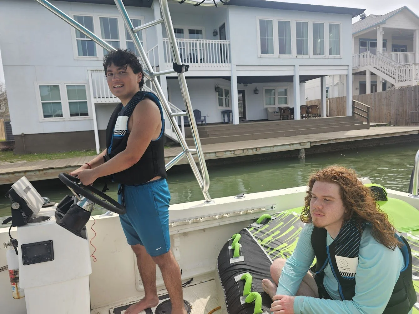 Two young men on a boat, one standing at the steering wheel and the other sitting, wearing life jackets, with a house and water in the background.