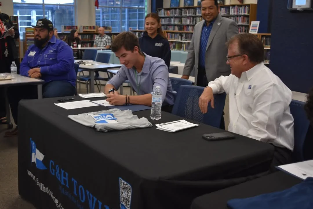 A young man signing a document at a table in a library, with several adults observing, including a man in a gray suit and a woman in a navy jacket.