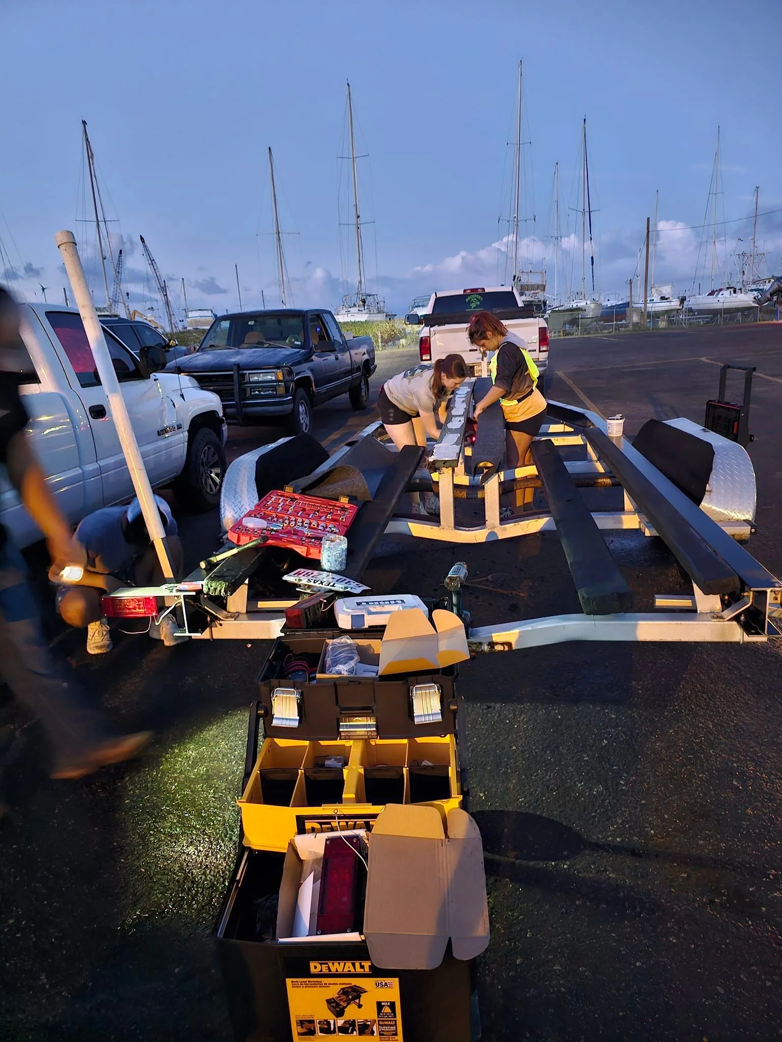 Two women are working on assembling a boat trailer in a parking lot near a marina with sailboats and cars in the background, at dusk.