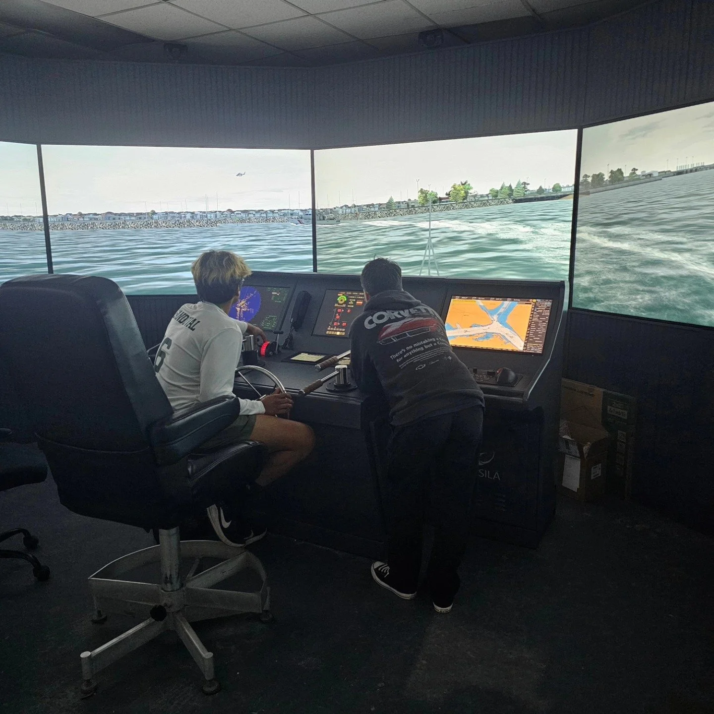 Two individuals operating a boat simulator with three large screens displaying water and shoreline views.