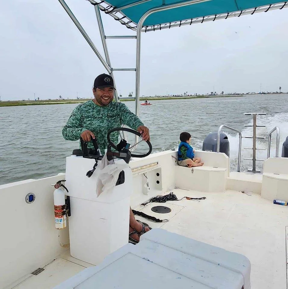A man steering a boat with a child sitting on the bench at the back, water and distant shoreline in the background.