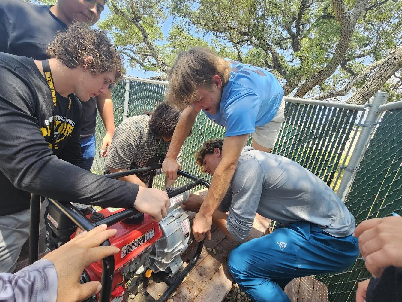 Group of young people working on a small portable generator outdoors, with a fenced area and trees in the background.