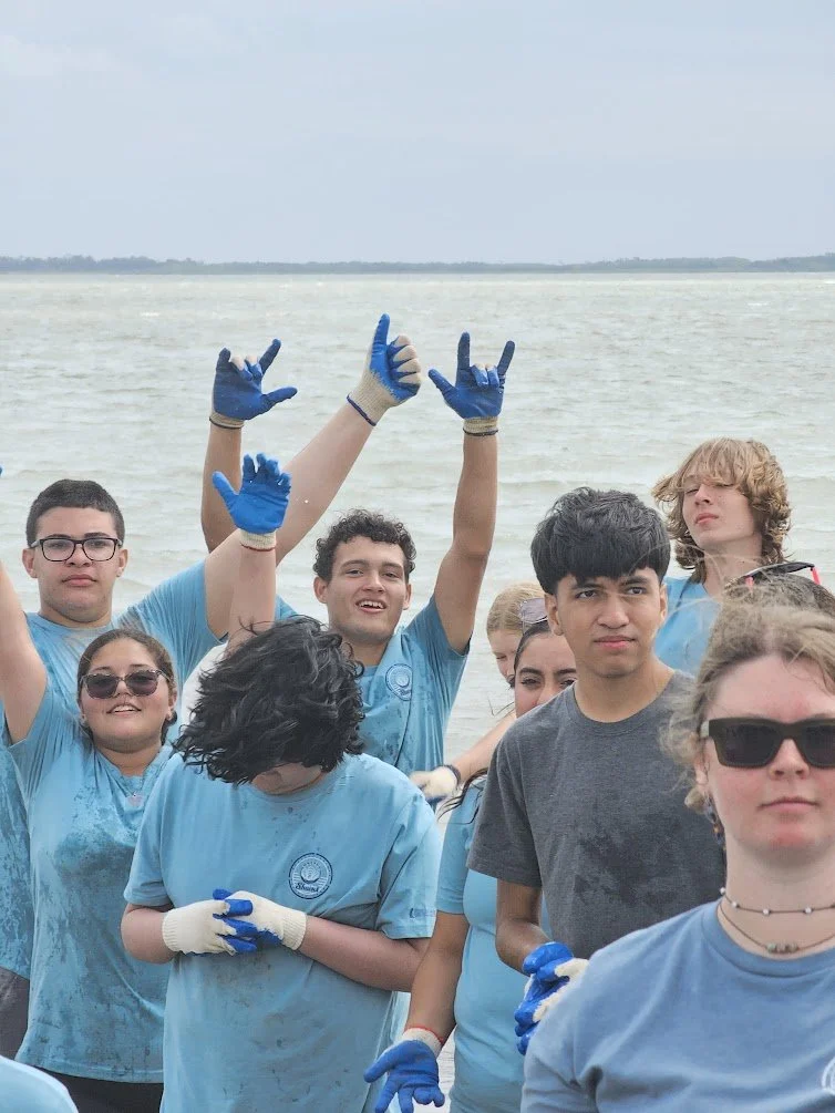 Group of teenagers wearing blue gloves and shirts near a body of water, some smiling and waving.