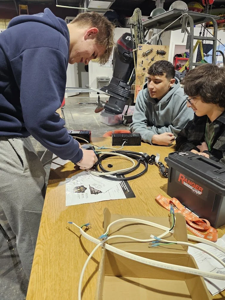 Three young men working together on an electrical project at a workshop table with tools, wiring, and instruction manual, in a garage or workshop setting.