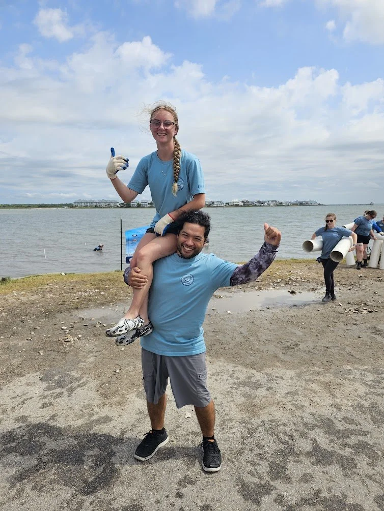 A man carrying a smiling woman on his shoulders at a waterfront during a group cleanup event. The woman is giving a thumbs-up, and other volunteers are in the background holding large pipes.