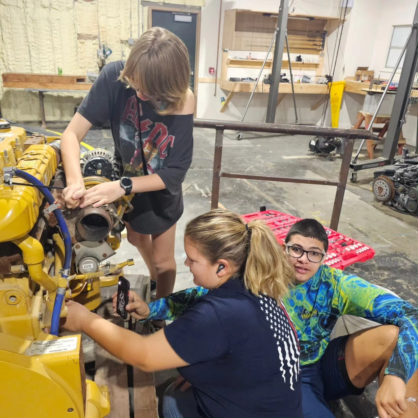 Three young people working on a large yellow mechanical engine in a workshop, with tools and equipment in the background.