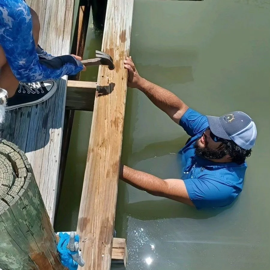 A man in a blue shirt, cap, and sunglasses is standing in water, holding a wooden dock with his hands, while a person in blue leggings and black sneakers is standing on the dock nearby.