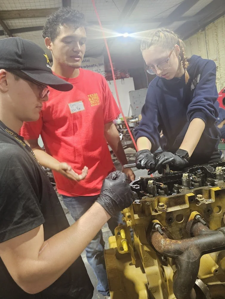 Three young adults working together on an engine in a workshop. One is operating a tool, while the others observe.