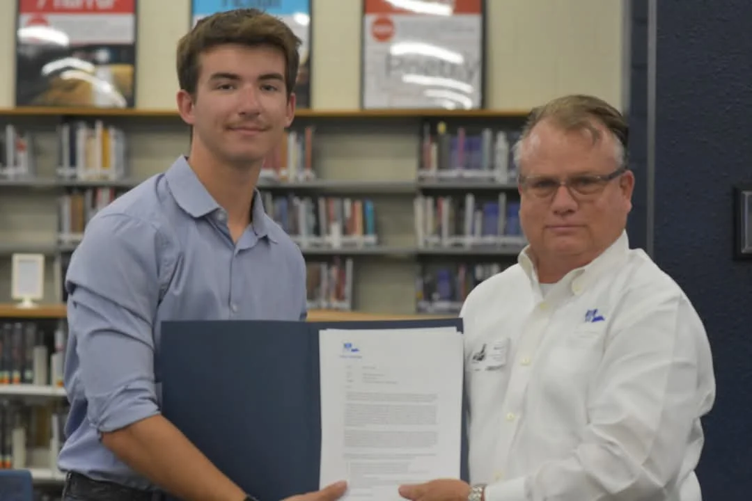A young man and an older man standing in a library, holding a large document together, smiling at the camera.