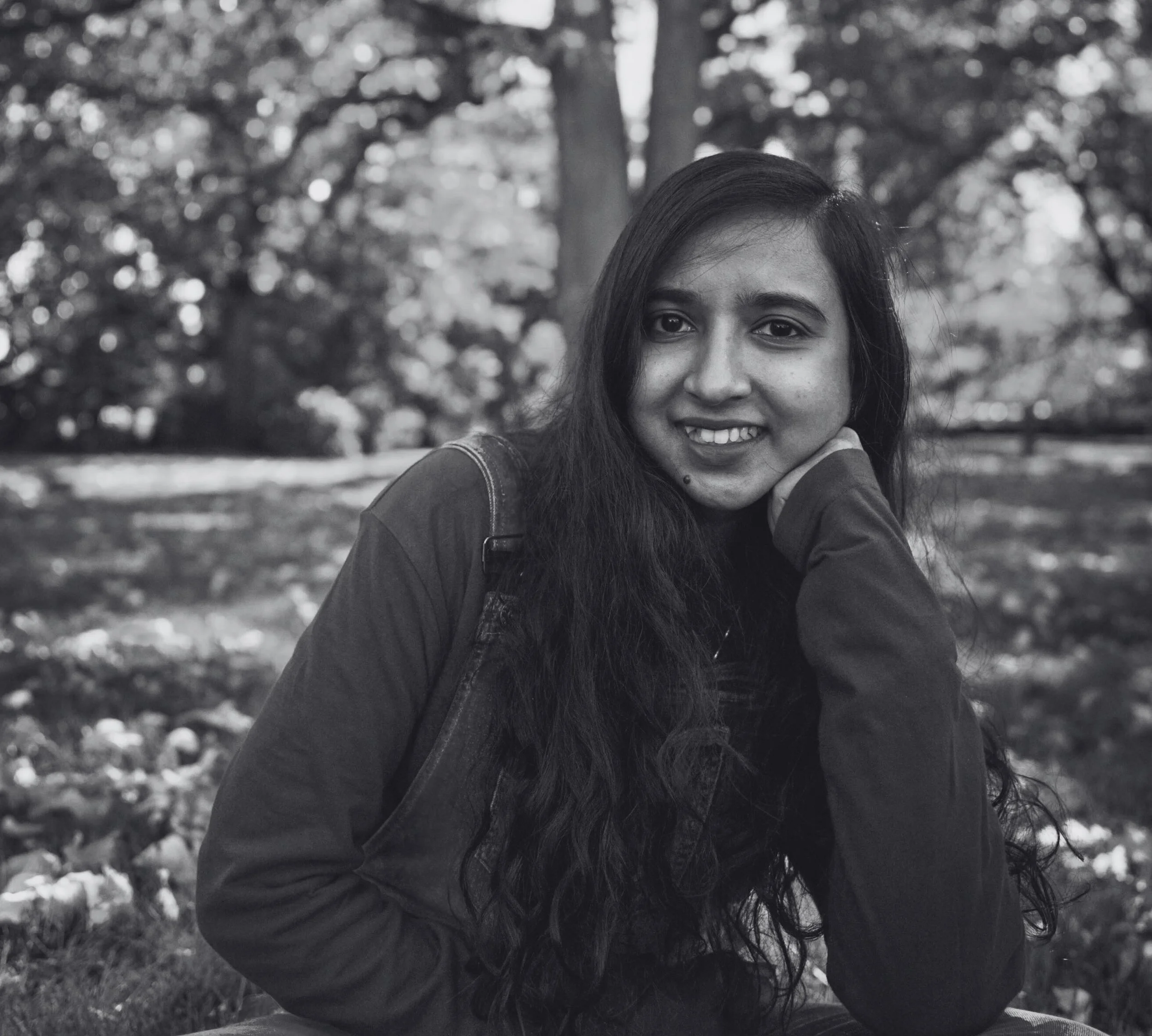 A smiling woman with long dark hair and a mole on her face, resting her chin on her hand outdoors in a park with trees in the background.