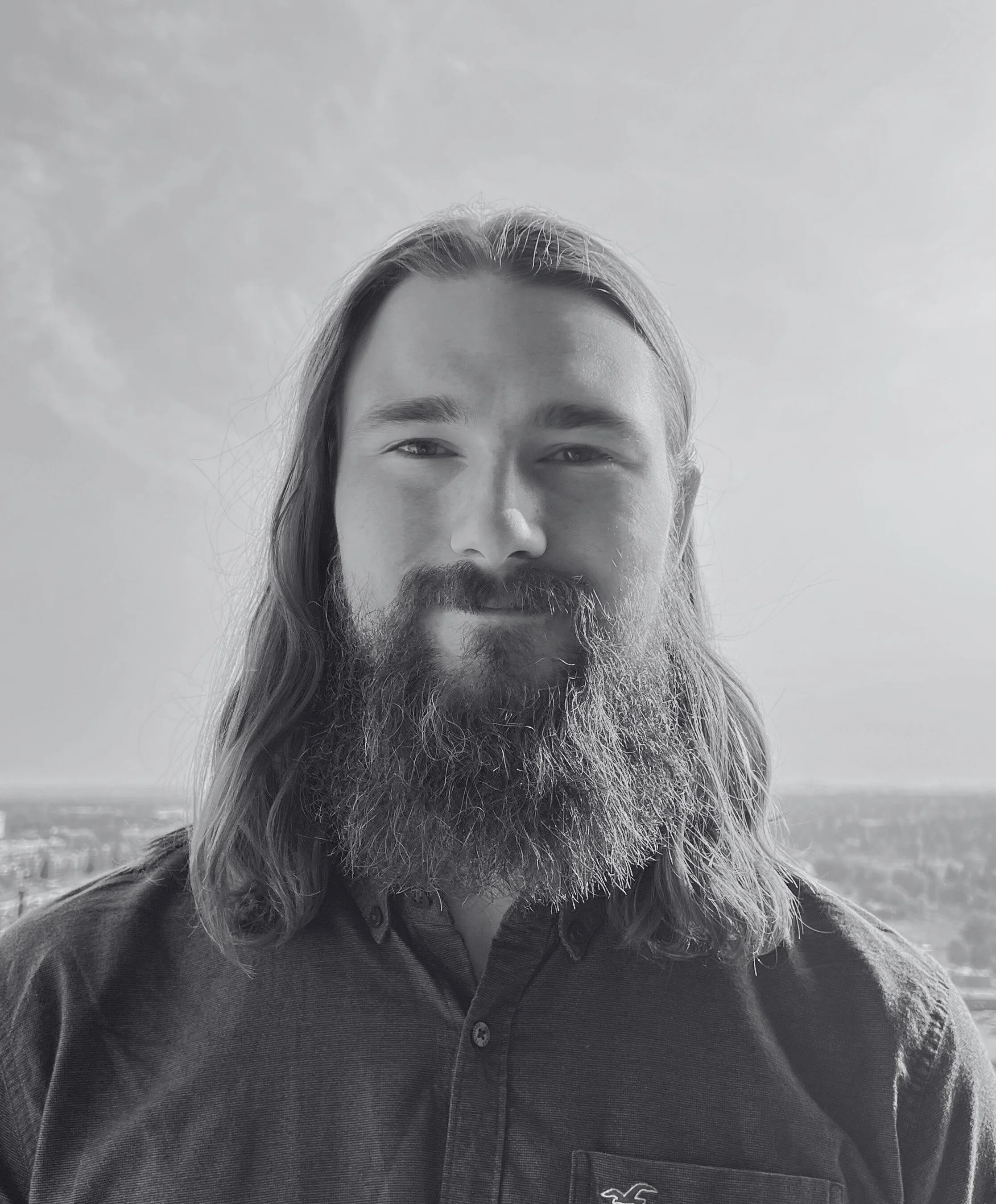 A man with long hair and a full beard smiling outdoors with a cityscape in the background, in black and white.
