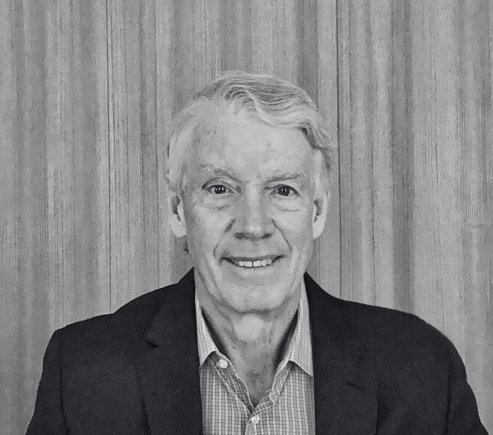 Black and white portrait of an older man wearing a suit jacket and collared shirt, smiling at camera, with a wooden background.