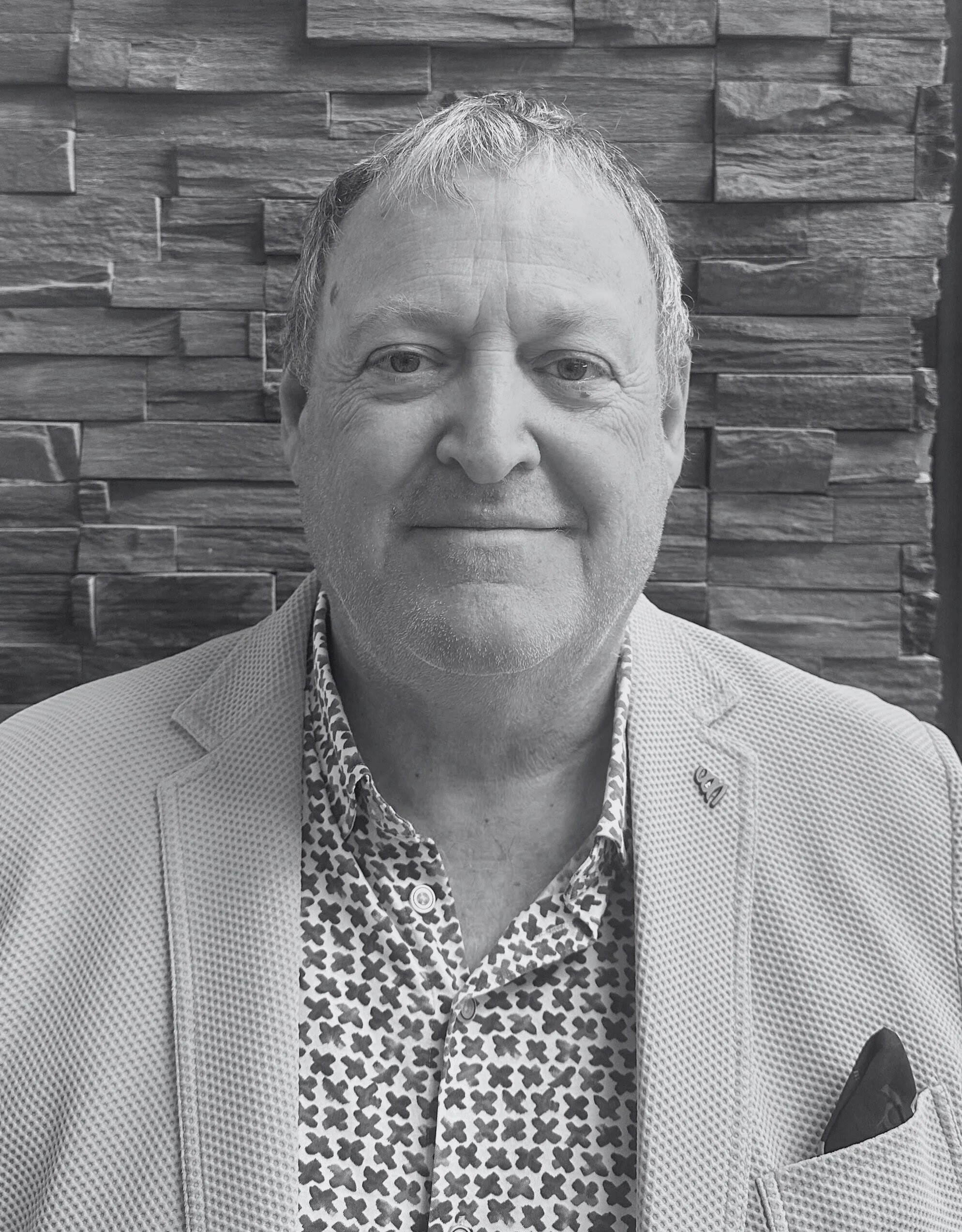 A middle-aged man with short hair and a slight smile, dressed in a patterned shirt and light-colored blazer, sitting in front of a textured stone wall.