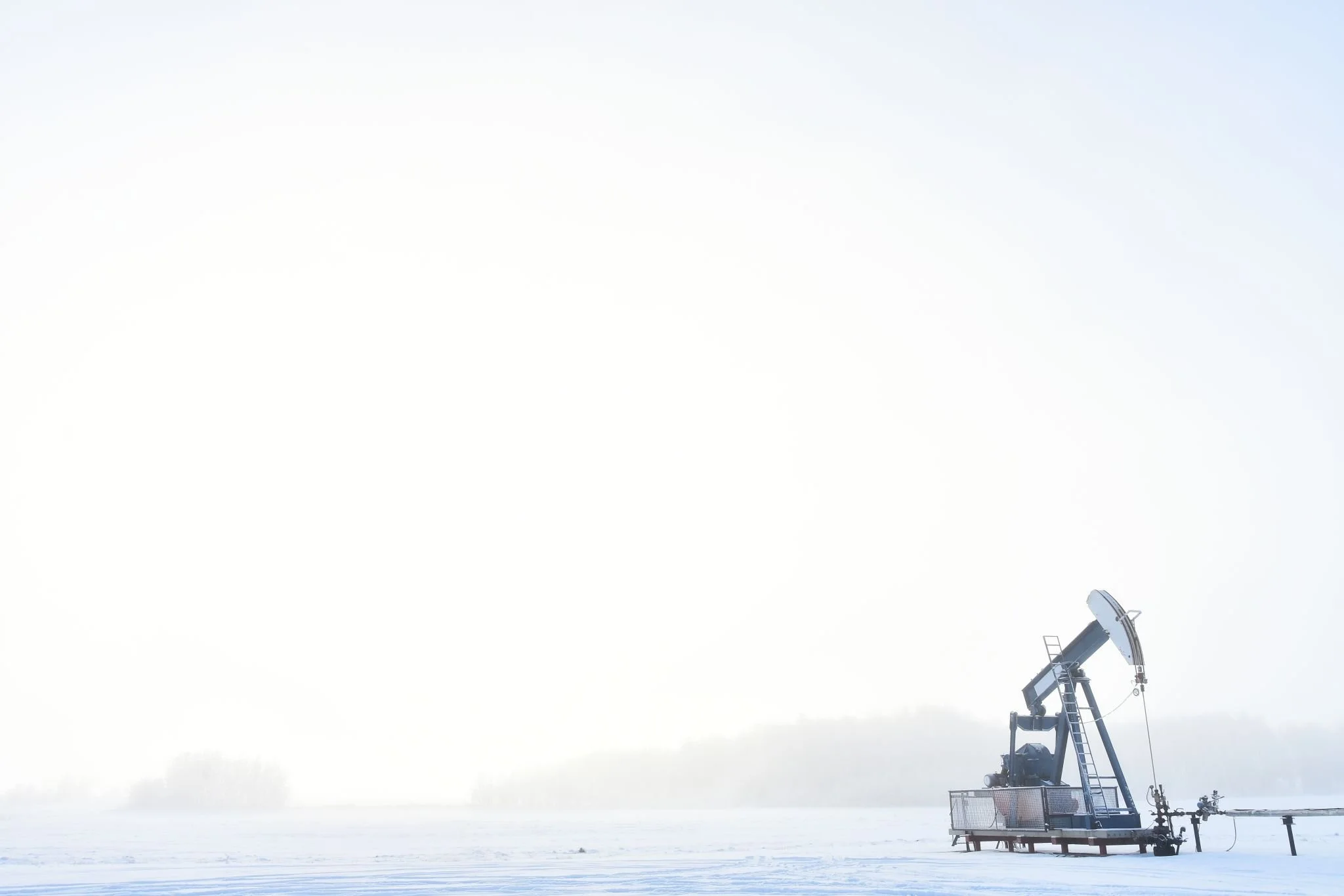 An oil pumpjack standing in a snowy landscape with overcast sky and distant hills.