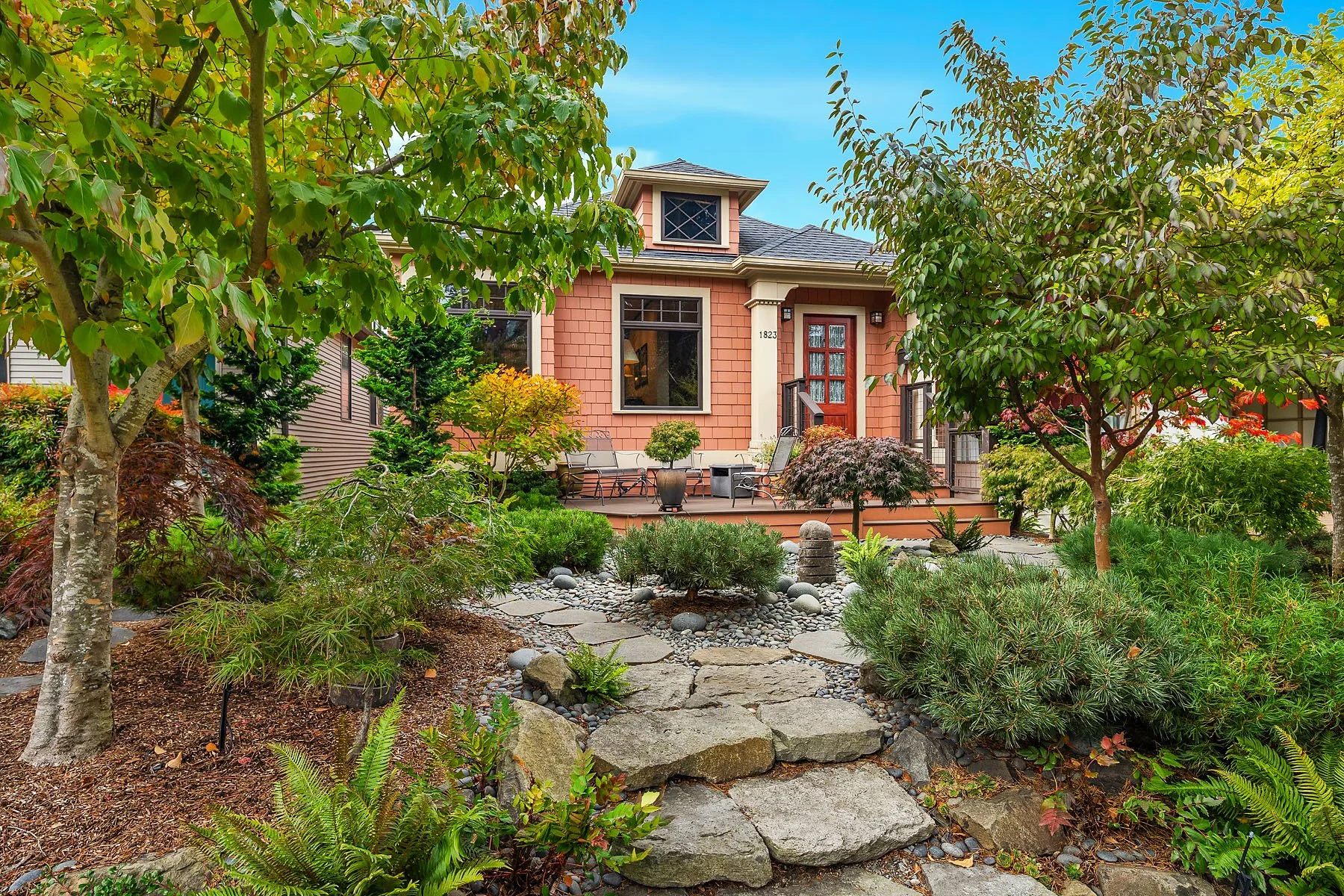 A vibrant landscaped garden in front of a pink craftsman house in Queen Anne neighborhood of Seattle with a deck. The garden features various Japanese maples, shrubs, small trees, ferns, and rocky pathways, with a mix of green and red foliage.