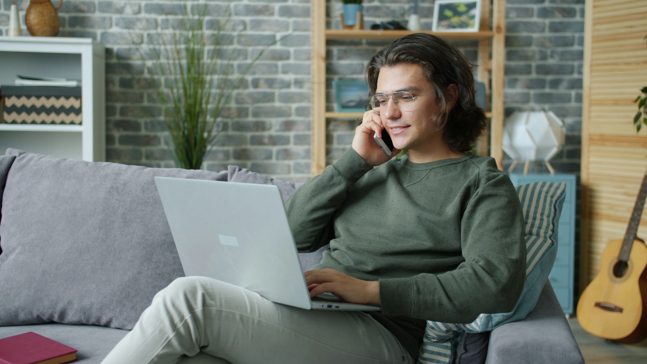 A young man sitting on a gray sofa, talking on a mobile phone, using a silver laptop, with a brick wall and wooden shelves in the background. There is an acoustic guitar on the right side of the image.