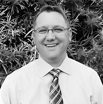 A smiling man wearing glasses, a white dress shirt, and a patterned tie standing outdoors with foliage in the background.