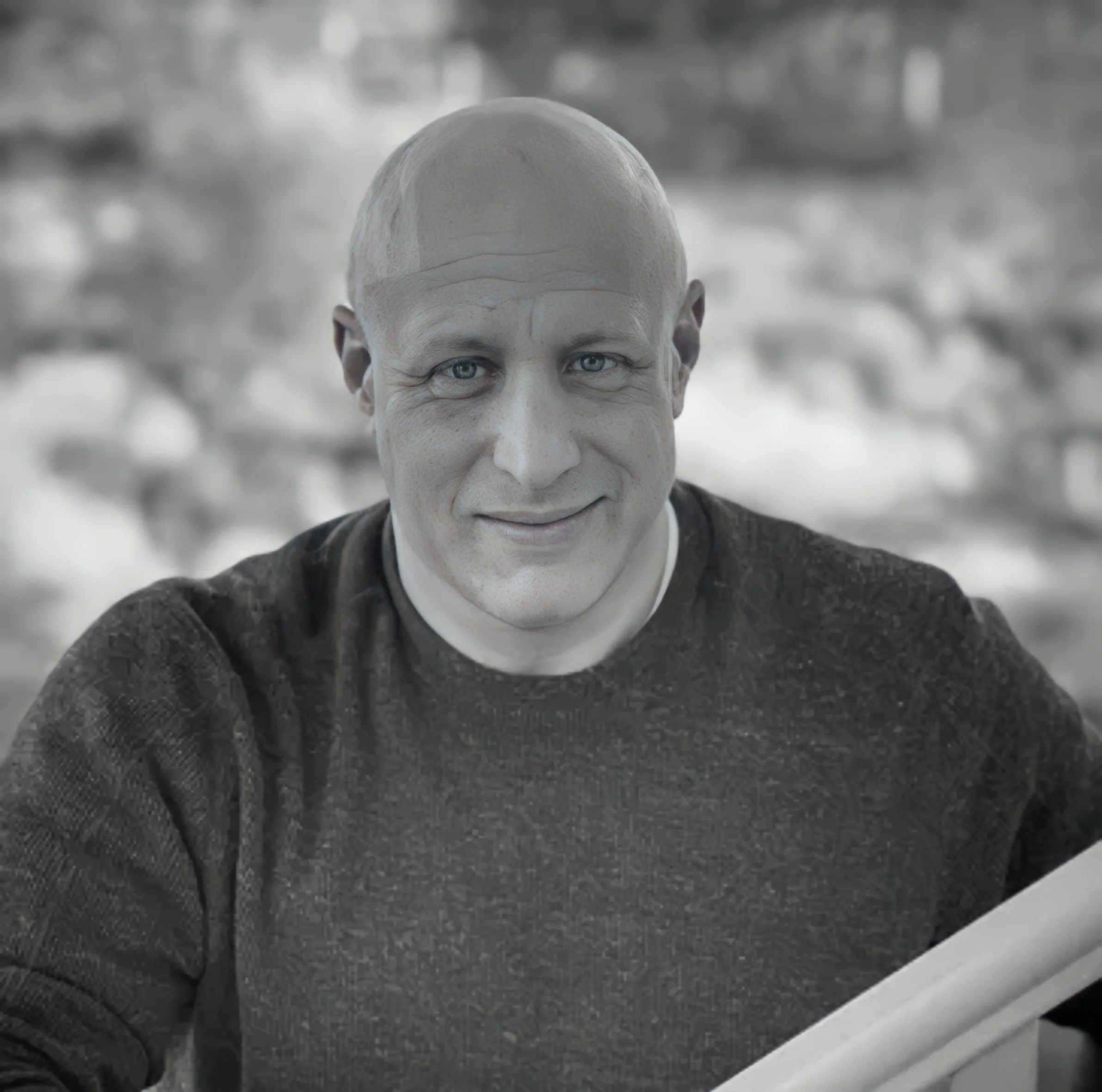 A man with a bald head, blue eyes, and a friendly smile, wearing a dark sweater, sitting outdoors with a blurred natural background.