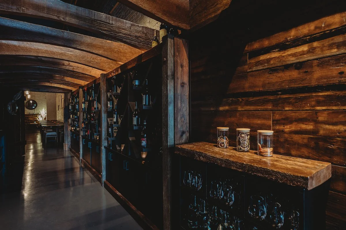 Interior of a dimly lit restaurant or bar with a wooden decor, featuring a wine or liquor bottle display and glass jars with decorative items on a wooden shelf.