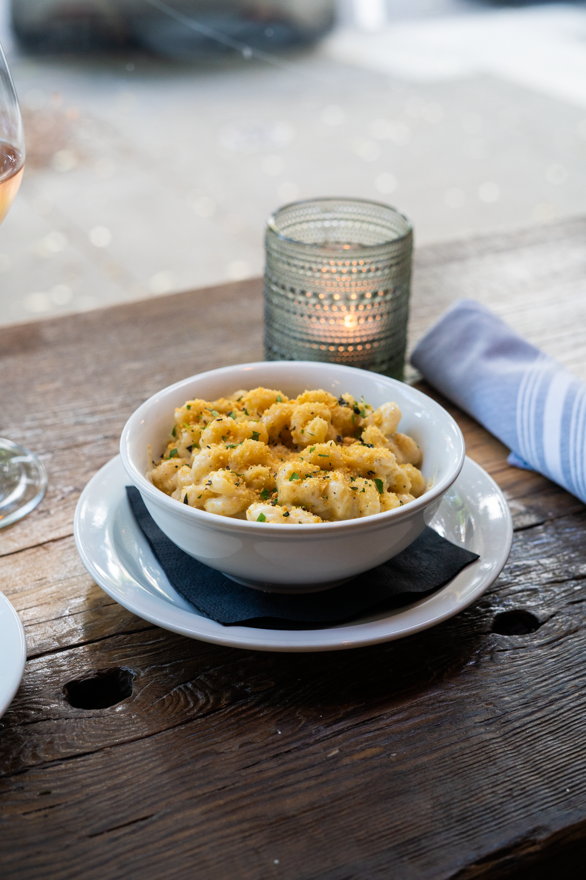 A bowl of macaroni and cheese on a wooden table with a candle in a glass holder, a blue and white striped napkin, and a glass of wine to the left.