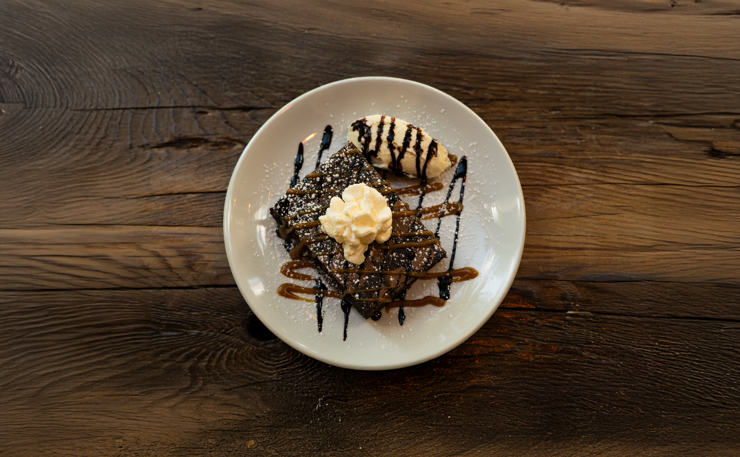 Chocolate brownie topped with whipped cream and chocolate drizzle, served with a side of vanilla ice cream with chocolate syrup, on a white plate.
