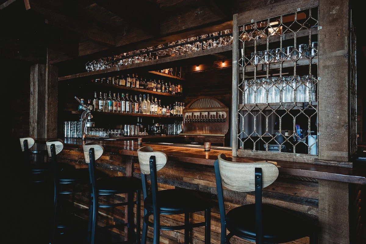 Interior of a dimly lit bar with a wooden counter, bottles of liquor on shelves, wine glasses stored overhead, and bar stools lined up.