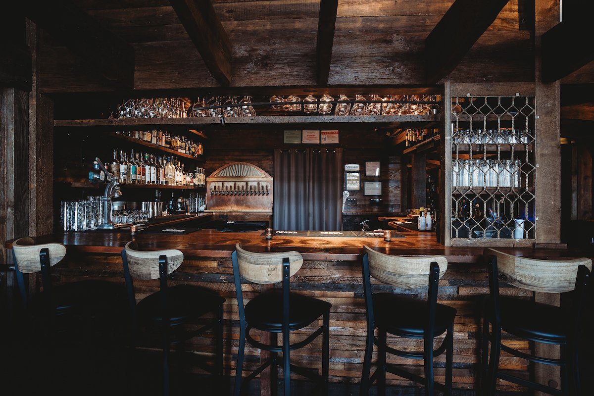 Empty bar with wooden counter, bar stools, and shelves with bottles and glasses in a dimly lit, rustic-style setting.