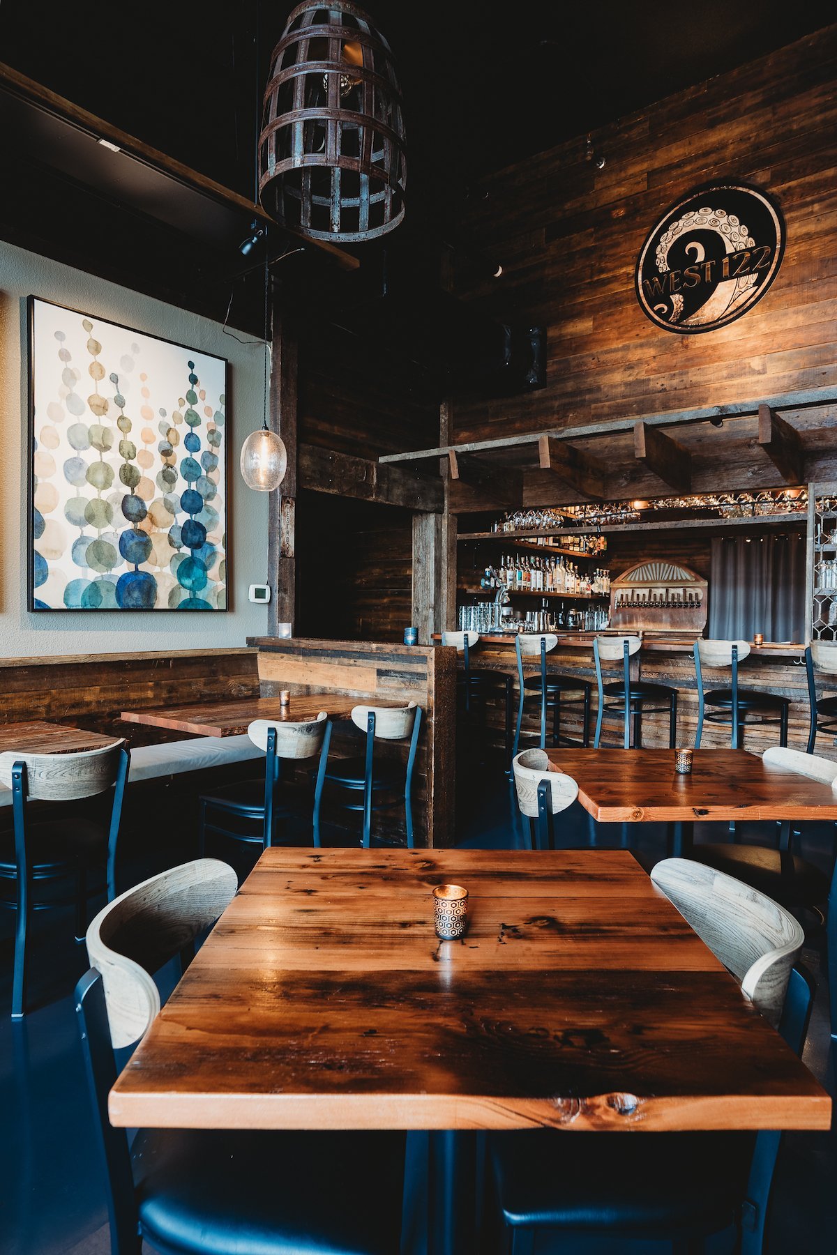 Interior of a rustic bar or restaurant with wooden tables, modern chairs, a bar counter with bottles, and decorative artwork on the wall.