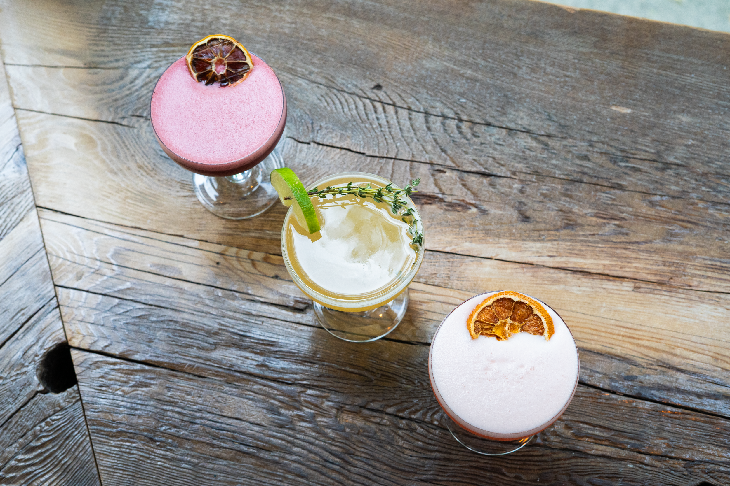 Three cocktails on a wooden table, each with a different garnish: a pink drink with a dried citrus slice, a clear yellow drink with a lime wedge and thyme sprig, and an orange-colored drink with a dried citrus slice.