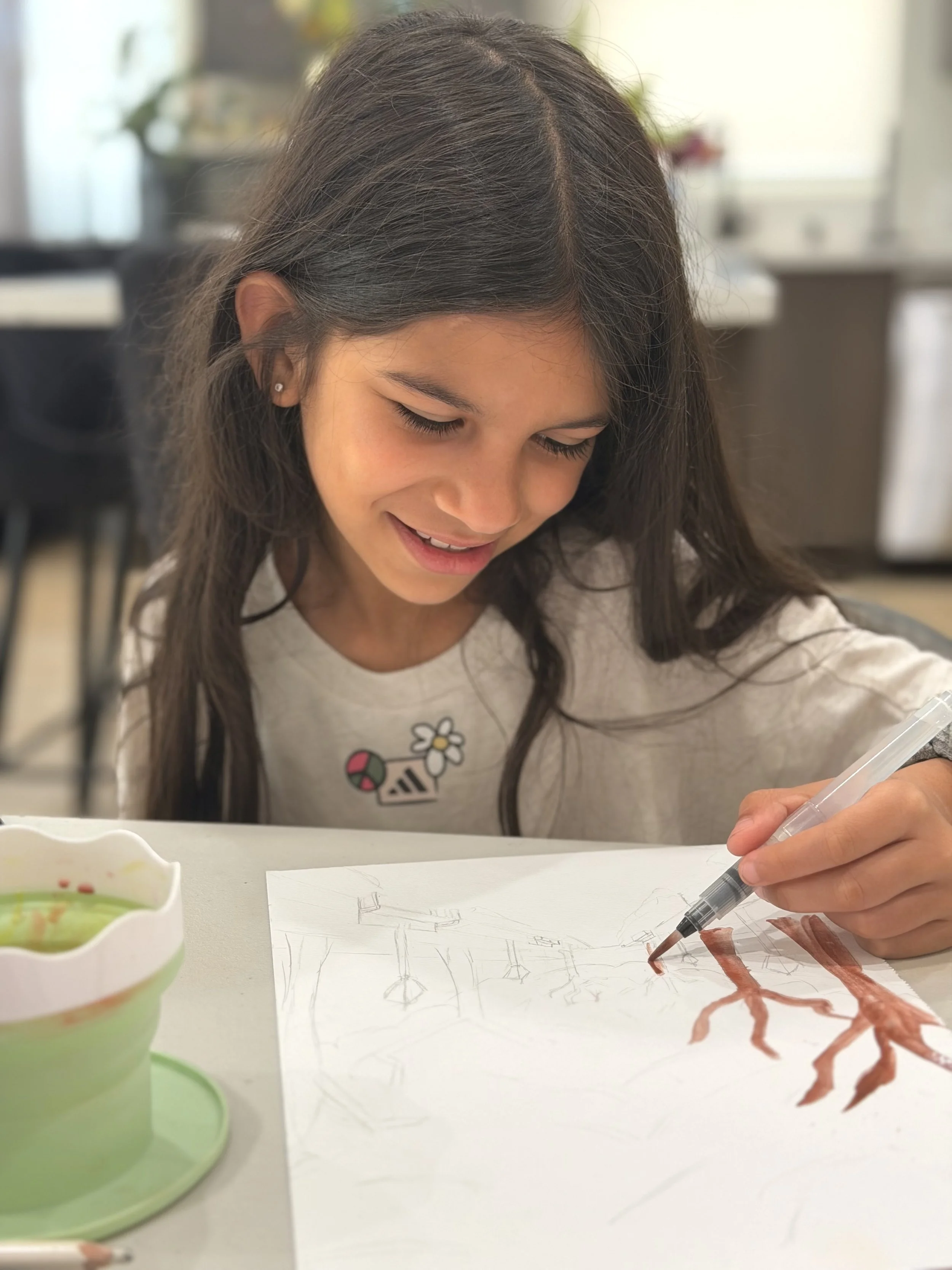 A girl drawing and coloring a tree on a sheet of paper at a table. She is smiling and appears to be enjoying her art activity in a bright room.
