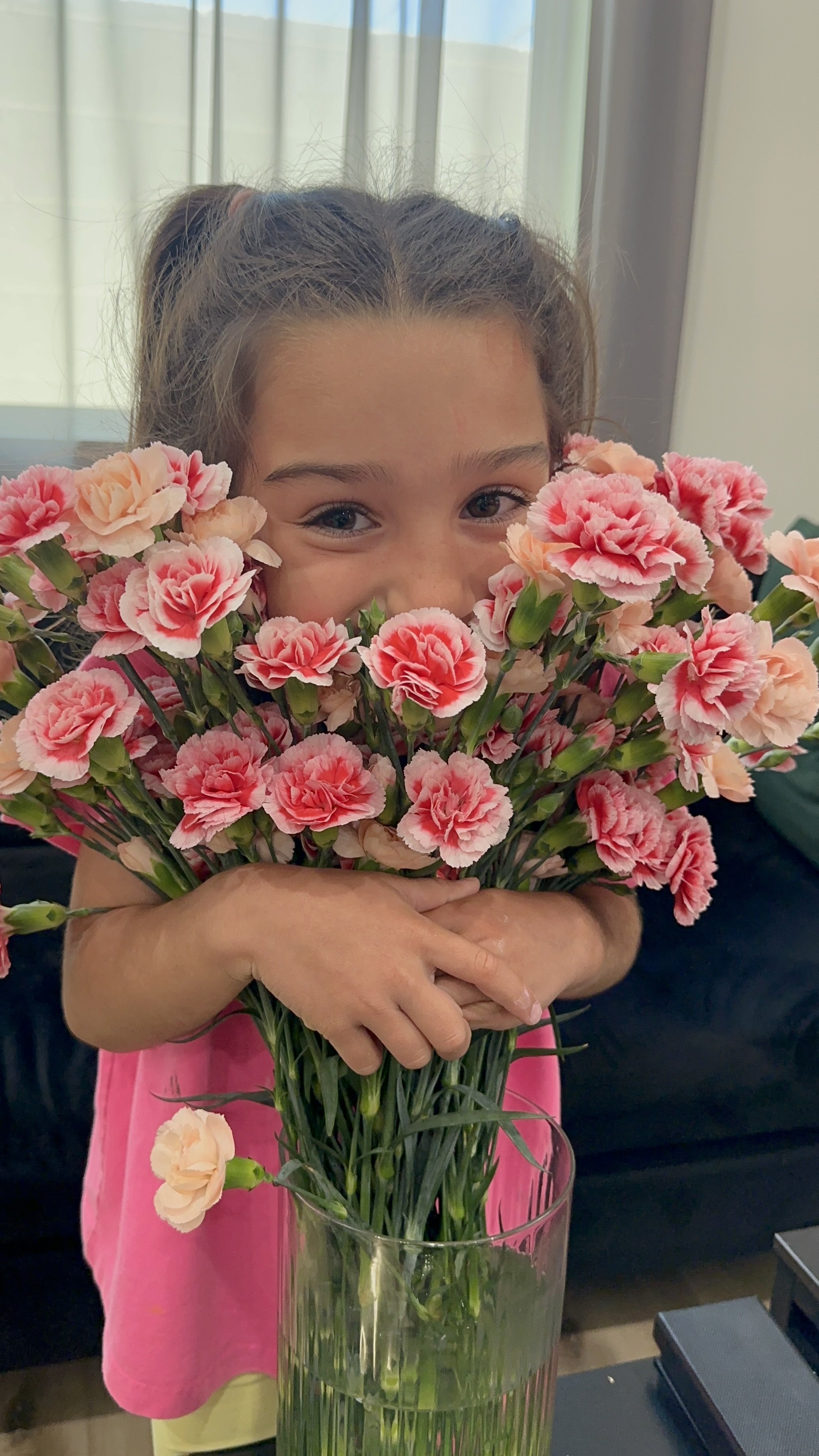 A young girl with brown hair holding a large bouquet of pink carnations in a glass vase in front of her face, with only her eyes and part of her forehead visible.