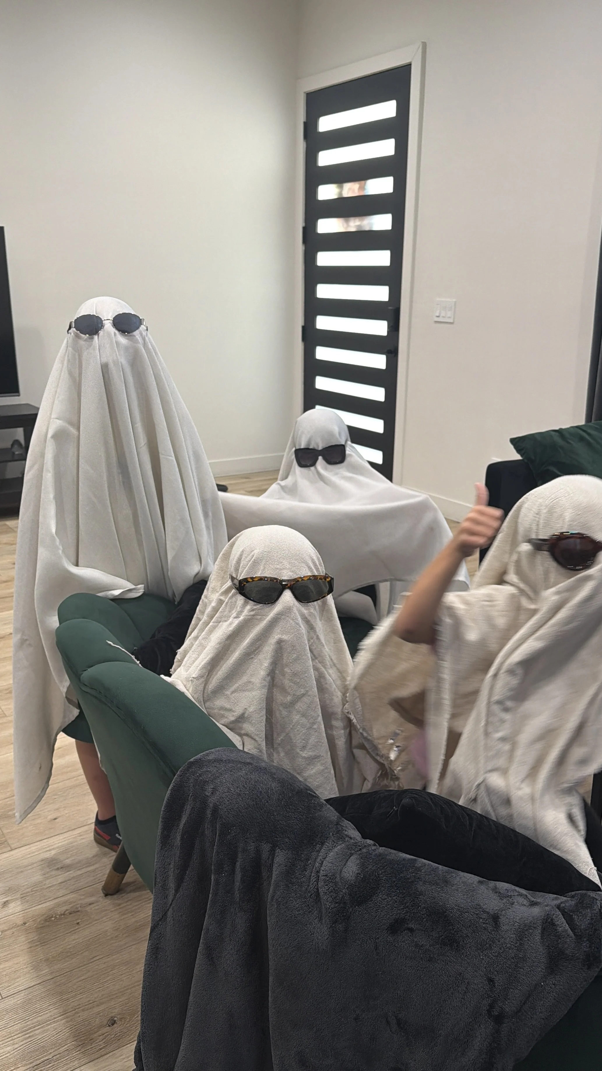People sitting on a couch and chair wearing white sheets over their heads with sunglasses, resembling ghosts.