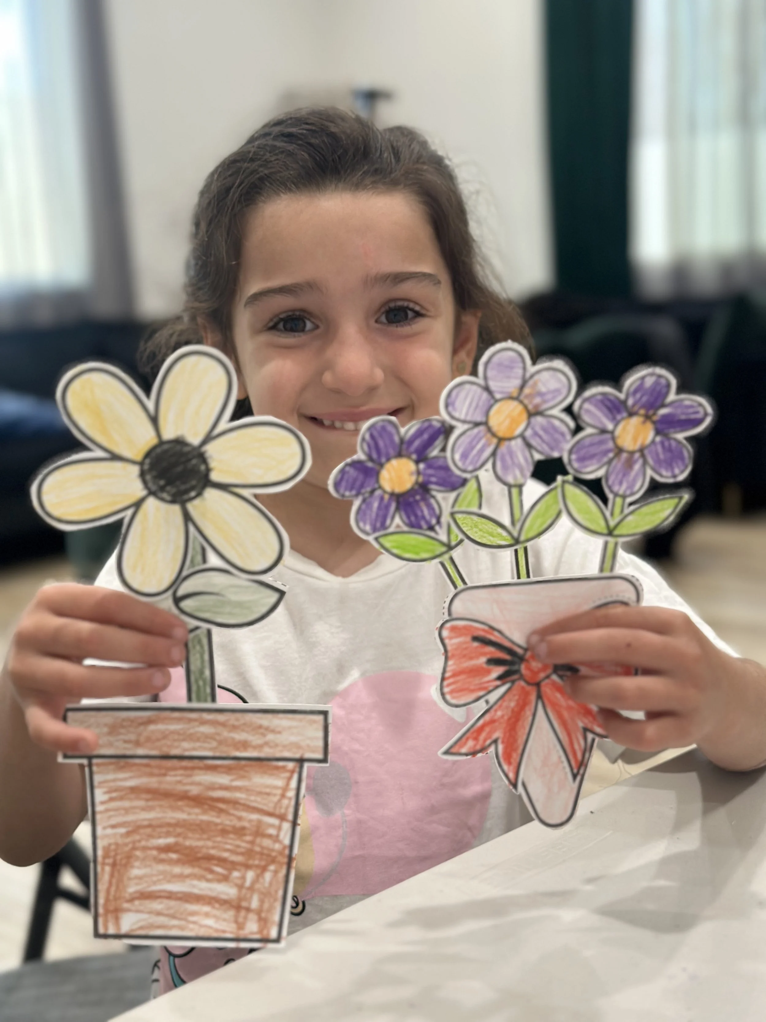 A young girl with curly brown hair holding hand-drawn and colored paper flowers, smiling at the camera.
