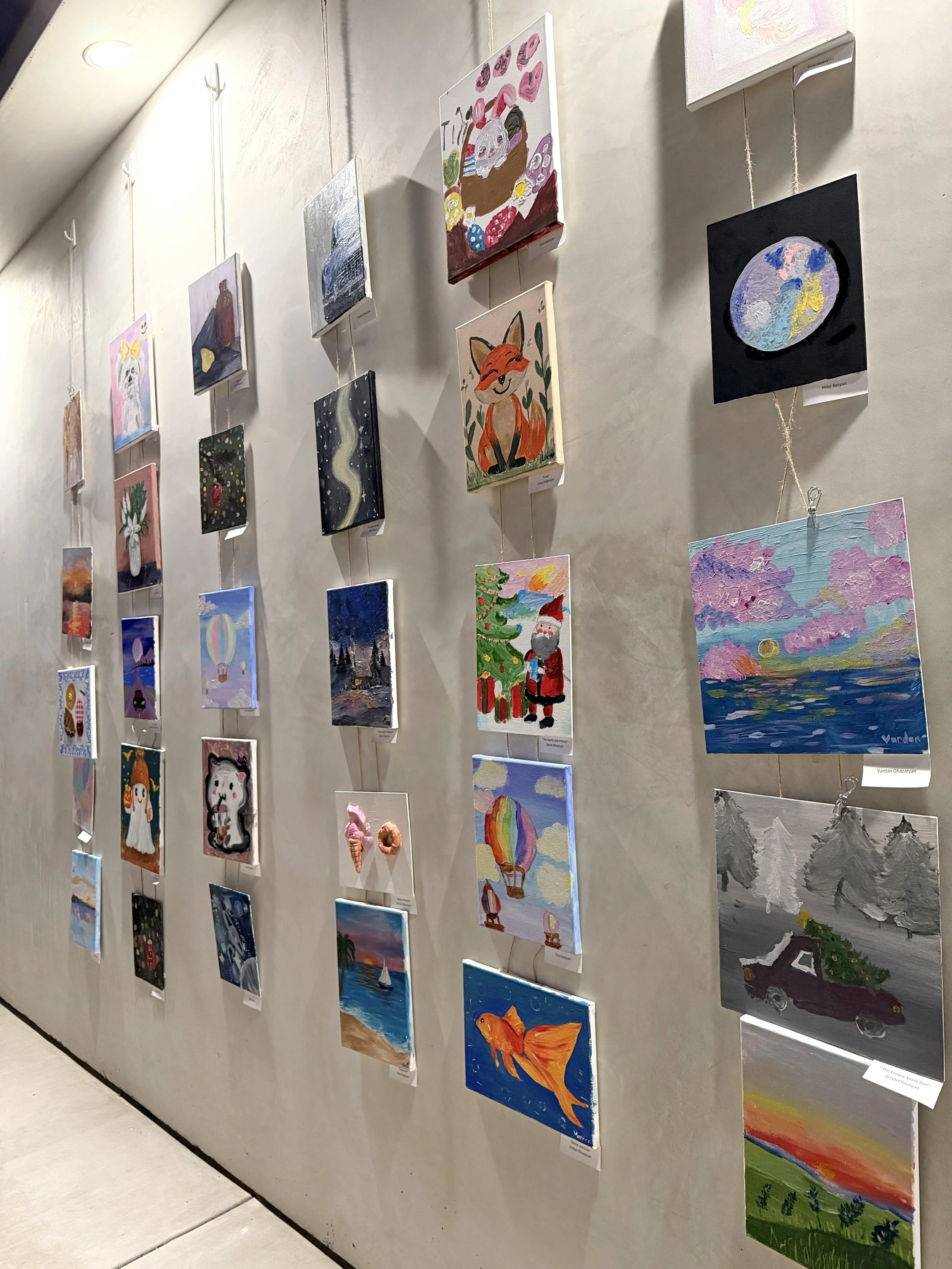 A gallery wall displaying landscape, animal, holiday, and abstract paintings, including a goldfish, a fox, a sunset, and a Christmas scene, arranged in rows and hanging on a beige wall.
