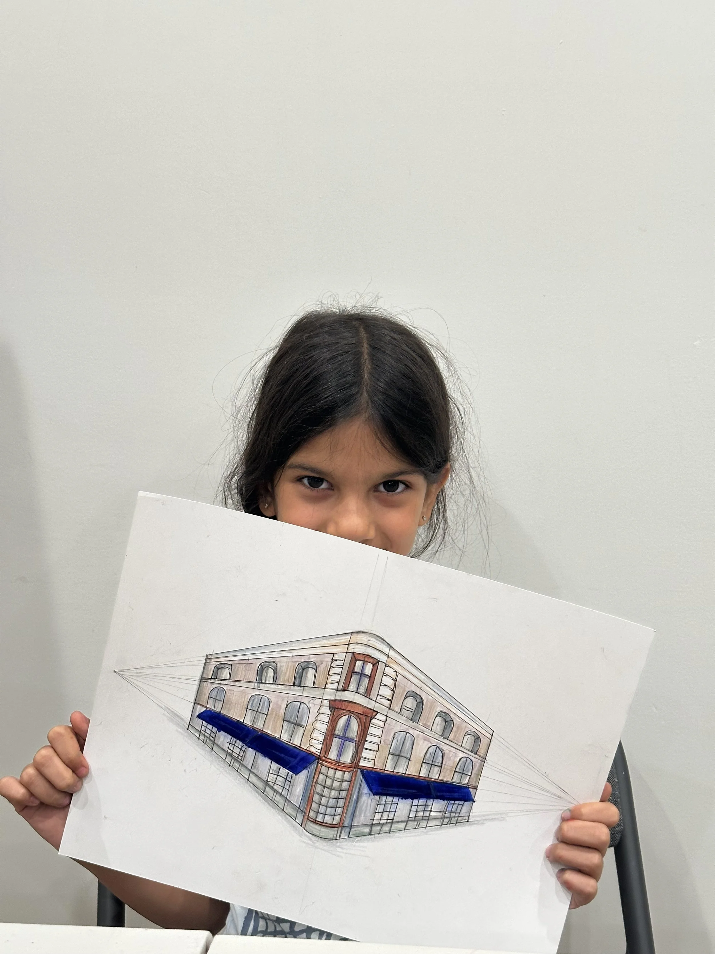 A young girl is holding a detailed architectural drawing of a building in front of her, partially covering her face, with only her eyes and forehead visible. The drawing includes windows, a corner turret, and a blue awning.
