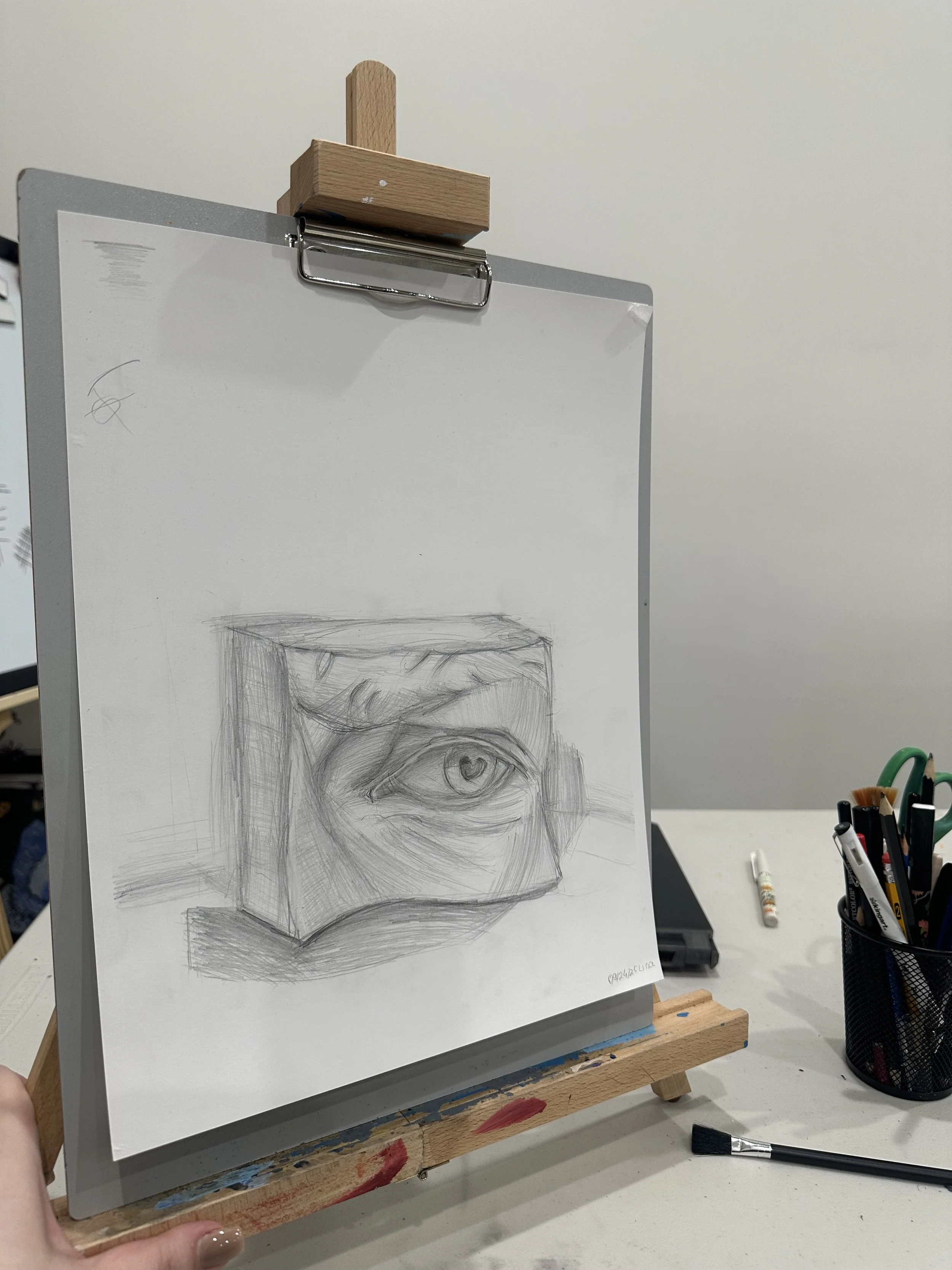A pencil sketch of a human eye on a small, rectangular piece of paper taped to a board, with art supplies visible on the table, all set in an art studio or classroom.