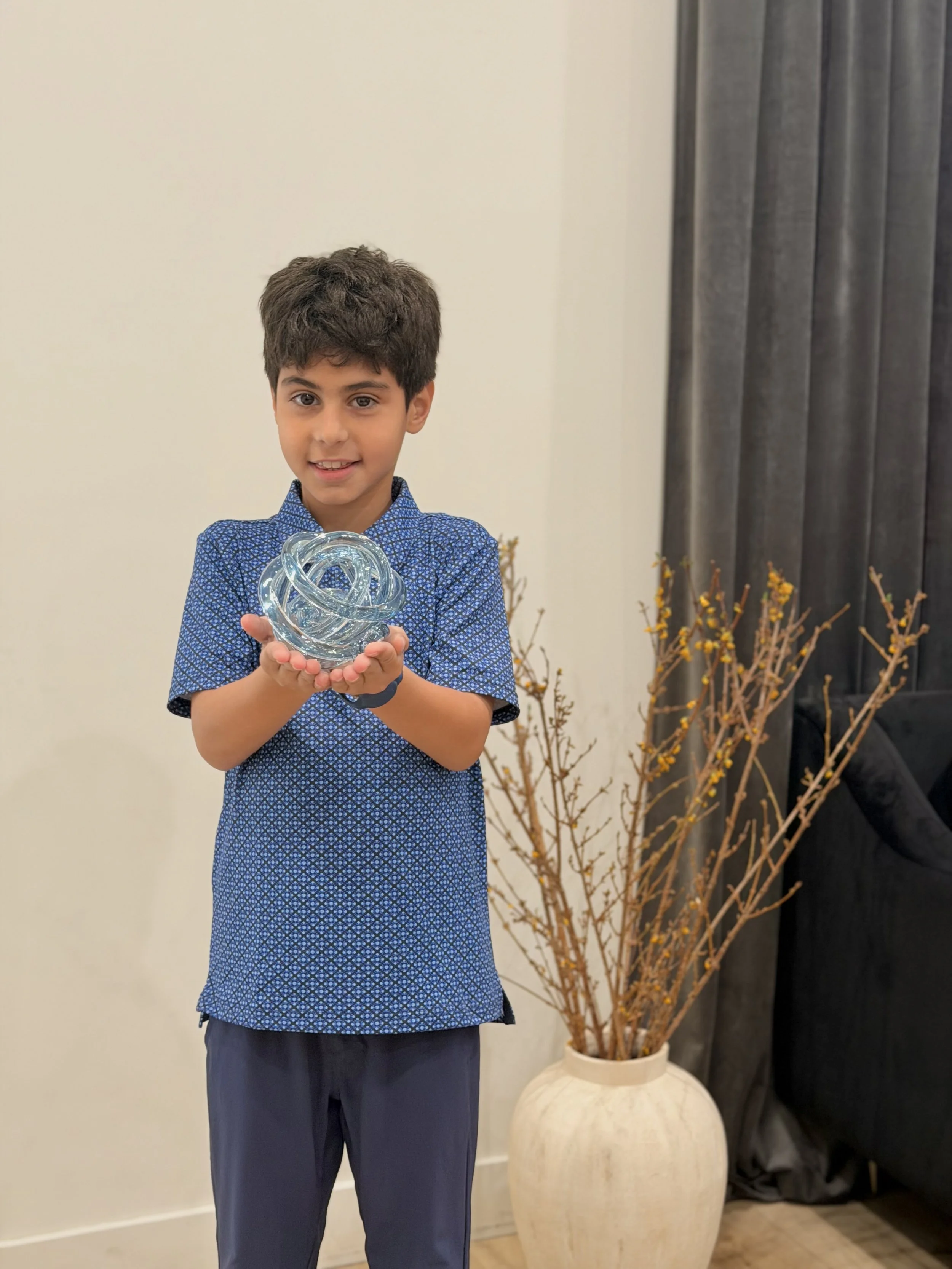 A young boy with dark hair wearing a blue patterned shirt and dark pants, standing indoors, holding a glass sculpture.