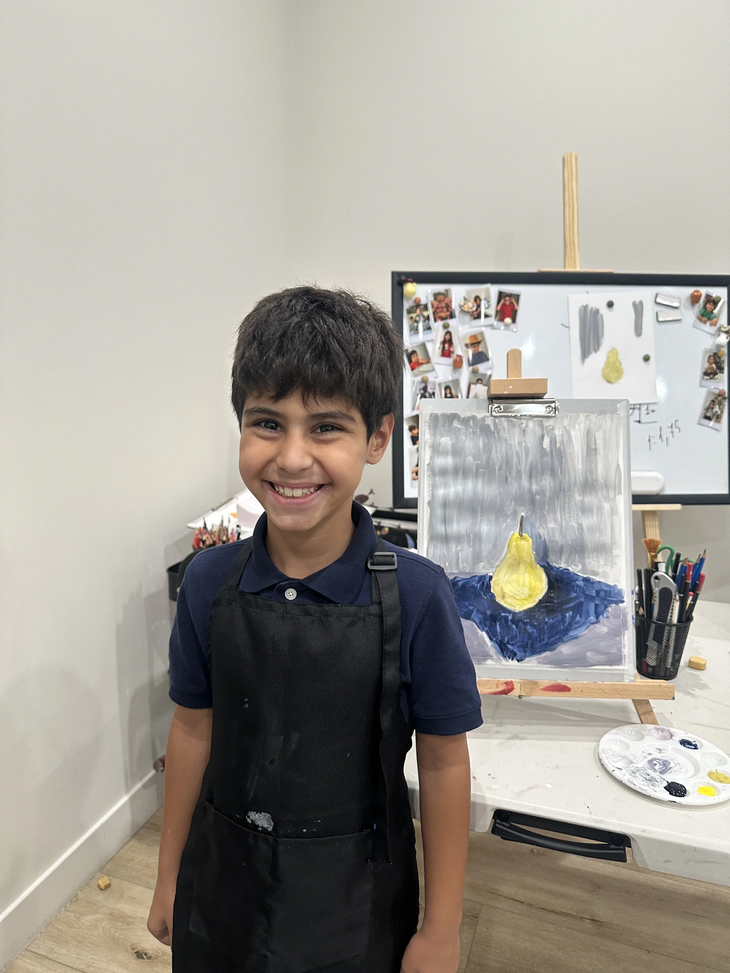 Smiling boy wearing a black apron standing in front of an easel with a painting of a yellow pear and in front of a table with painting supplies.