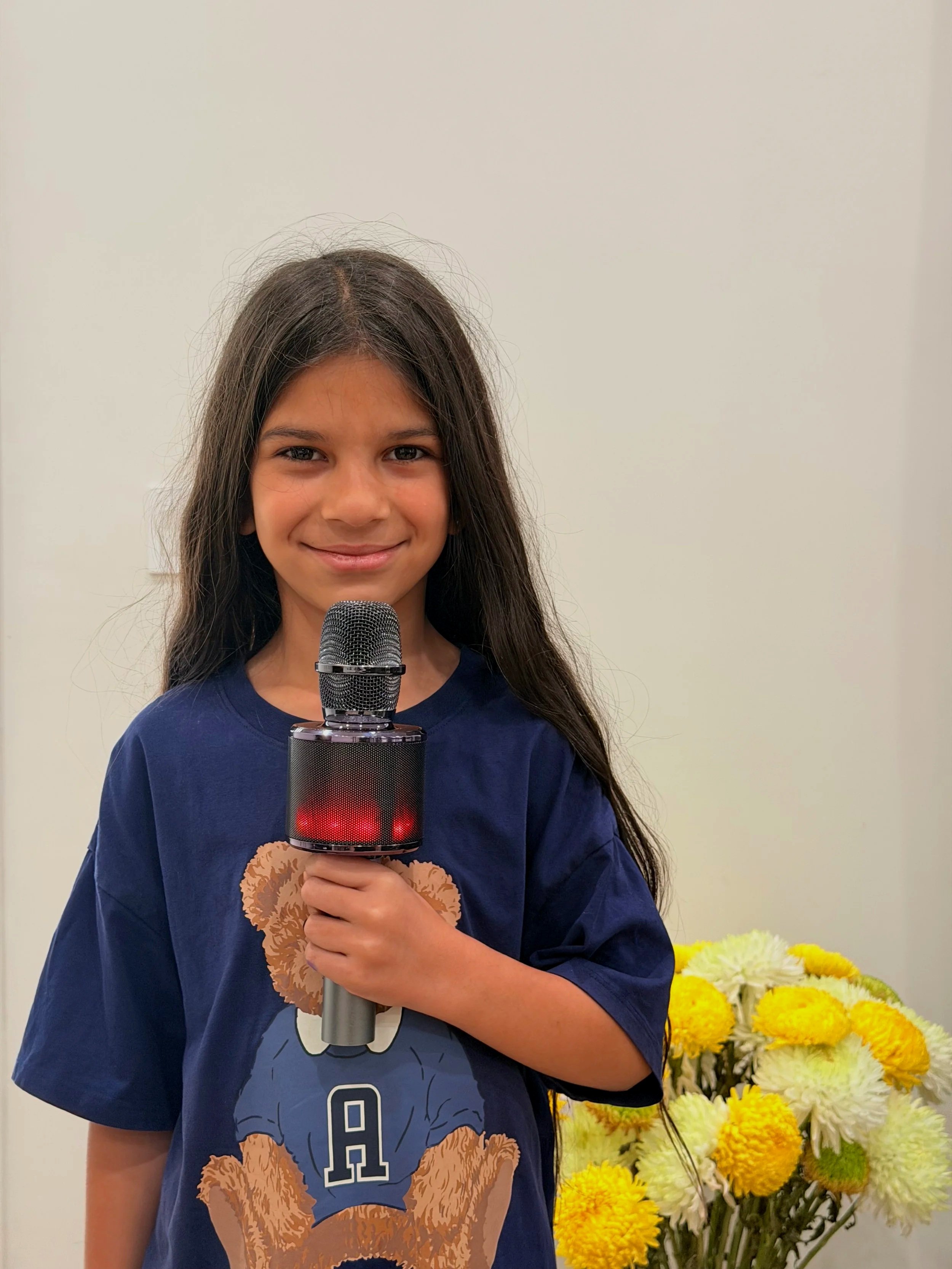 A young girl with long dark hair smiling as she holds a microphone, wearing a navy blue T-shirt with a bear graphic and the letter 'A' on it. Behind her is a bouquet of yellow and white flowers against a plain, light-colored wall.