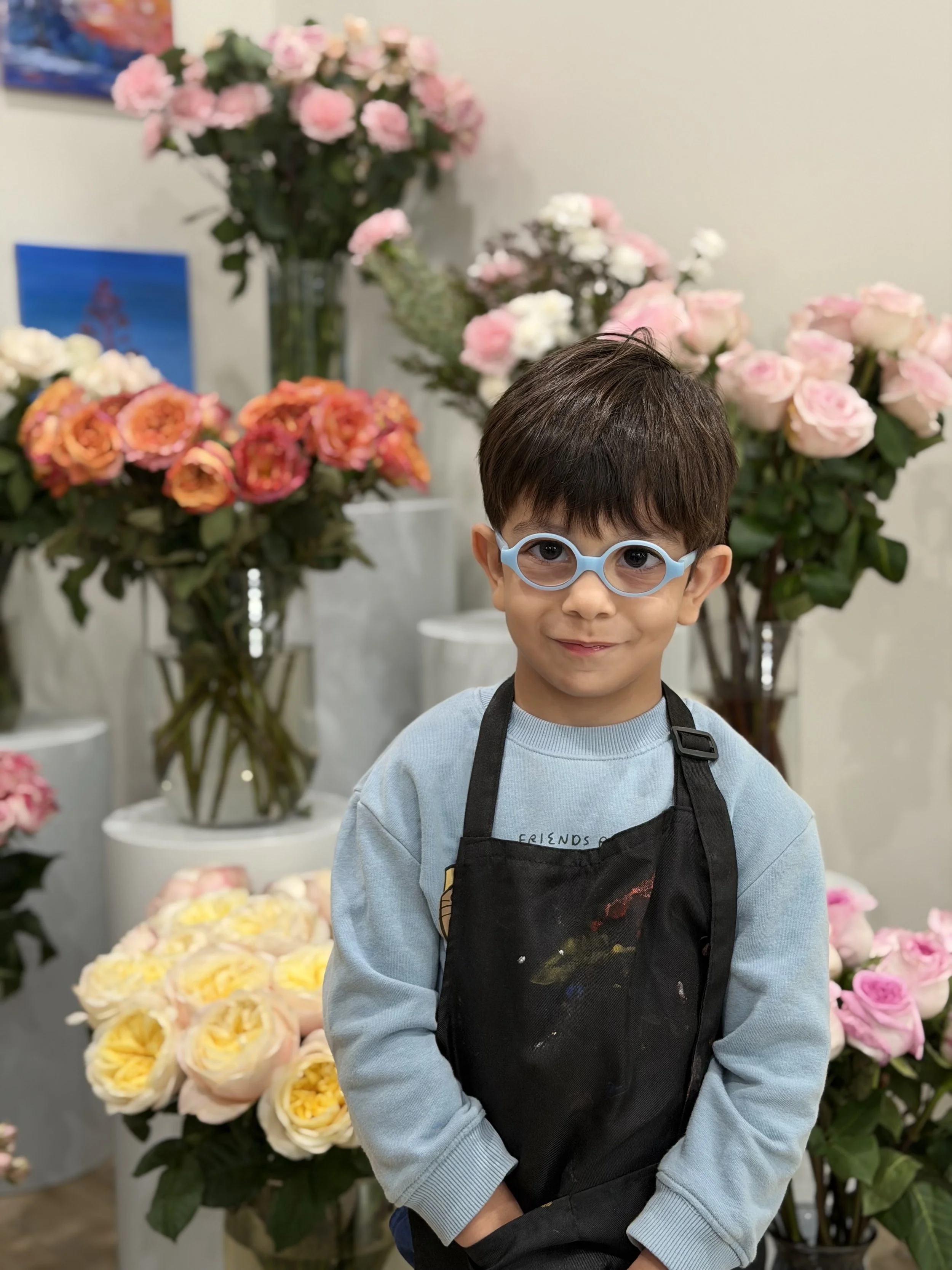 Young boy wearing blue glasses and a black apron standing in front of colorful flower arrangements in vases.