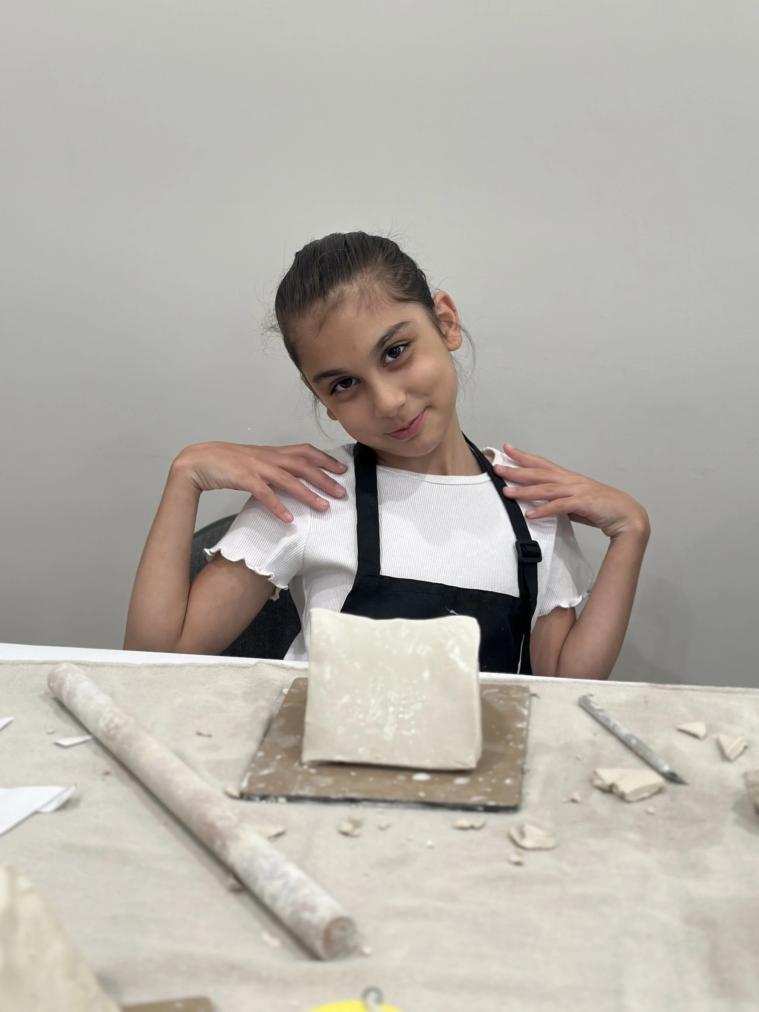 Young girl with dark hair in a bun, wearing a white top with ruffled sleeves and a black apron, sitting at a table with plaster or clay, hands near her shoulders, posing for the photo against a plain white wall.