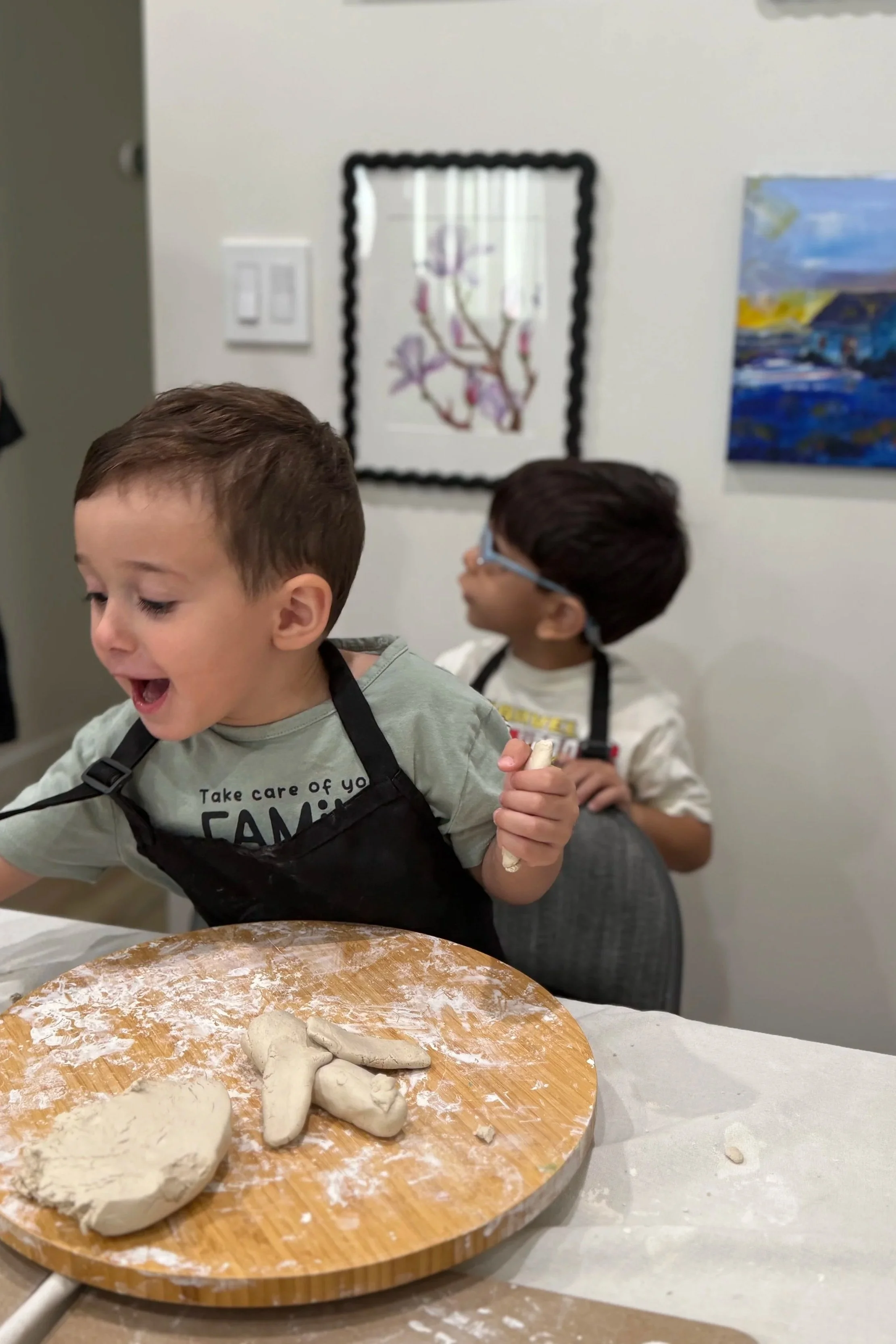 Two young boys are engaging in a cookie or clay crafting activity. One boy is smiling and holding a piece of dough while the other boy is wearing glasses and focused on their project. On the table, there is a wooden board with shaped dough pieces, an