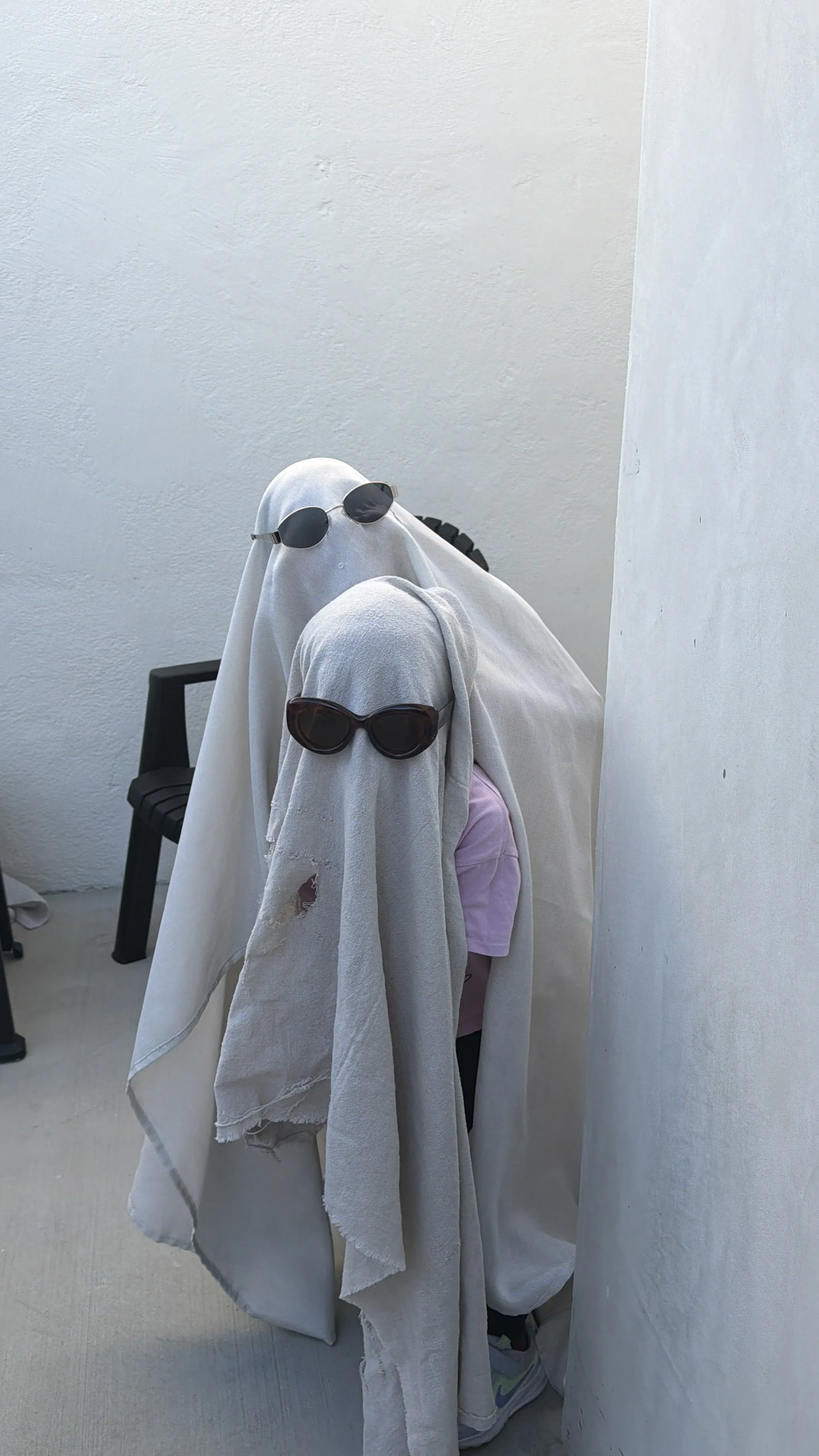 A person dressed as a ghost with a white sheet over their head, wearing sunglasses, and hiding behind a white curtain. They are in a room with white walls and a black chair in the background.