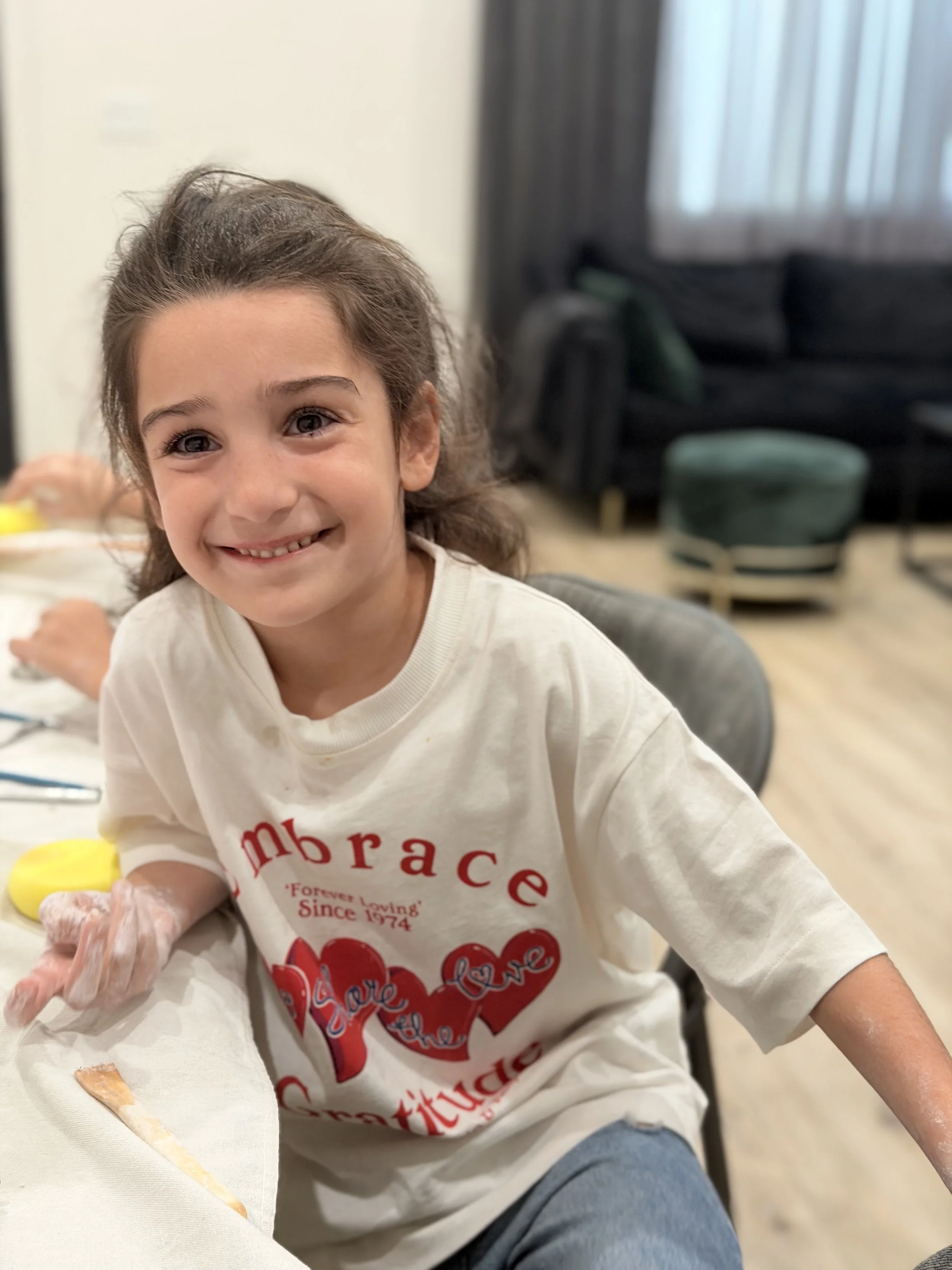 A smiling young girl with brown hair, wearing a white t-shirt with red text, sitting at a table with a white tablecloth, and some food items, in an indoor setting with a dark curtain and window in the background.