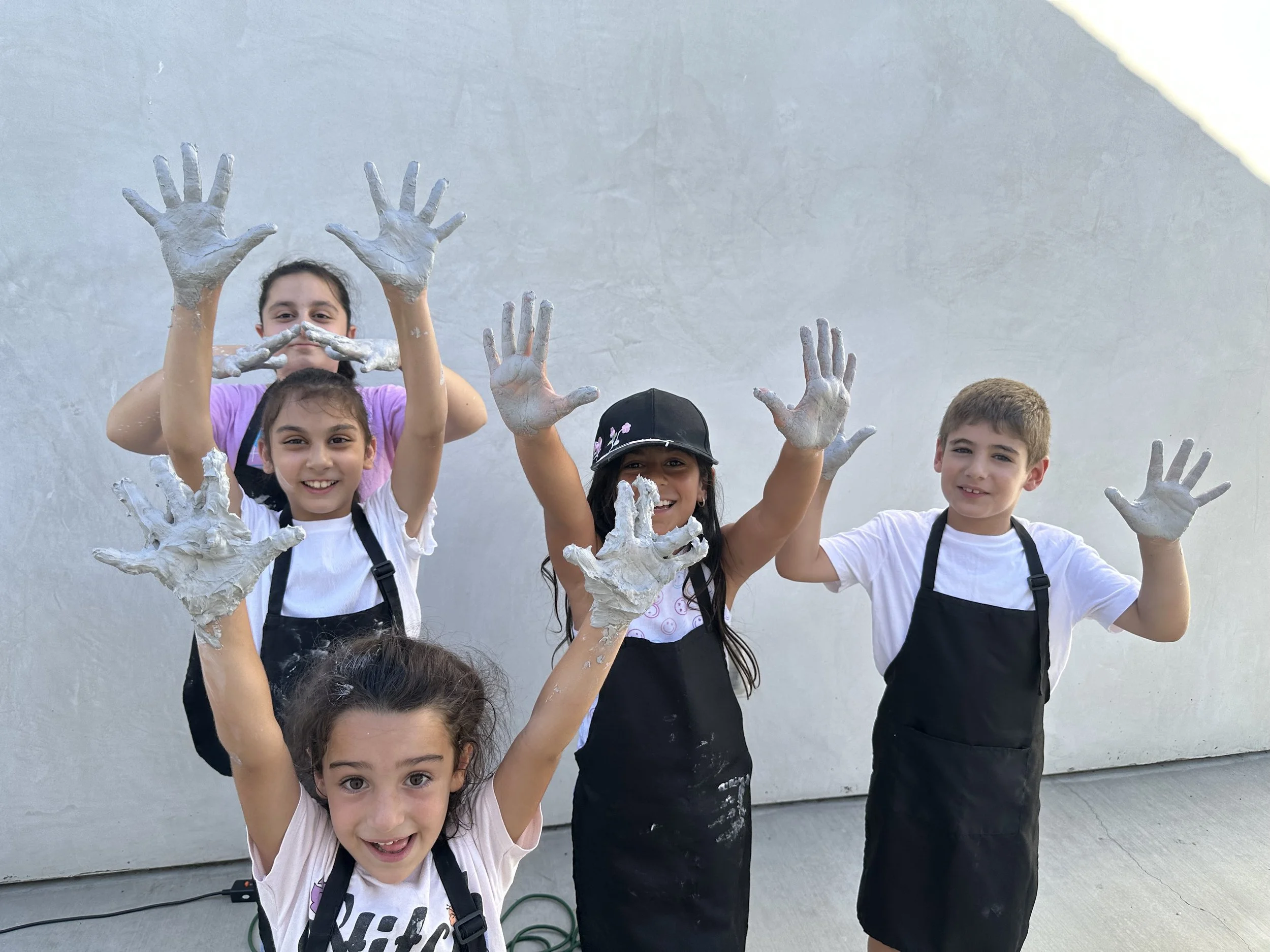 Group of children with painted hands raising their hands and smiling outdoors against a plain wall.