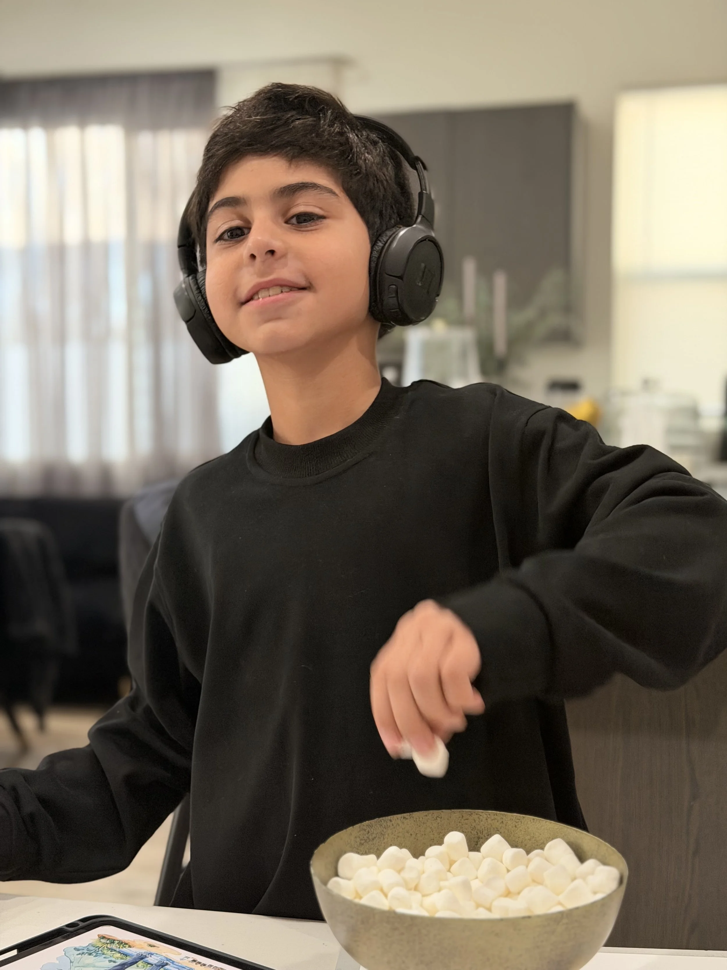 A young boy with dark hair and wearing a black long-sleeve shirt, sitting at a table, wearing large black headphones, and smiling slightly. There is a large bowl of marshmallows in front of him, and a gaming tablet and a smartphone on the table. The 