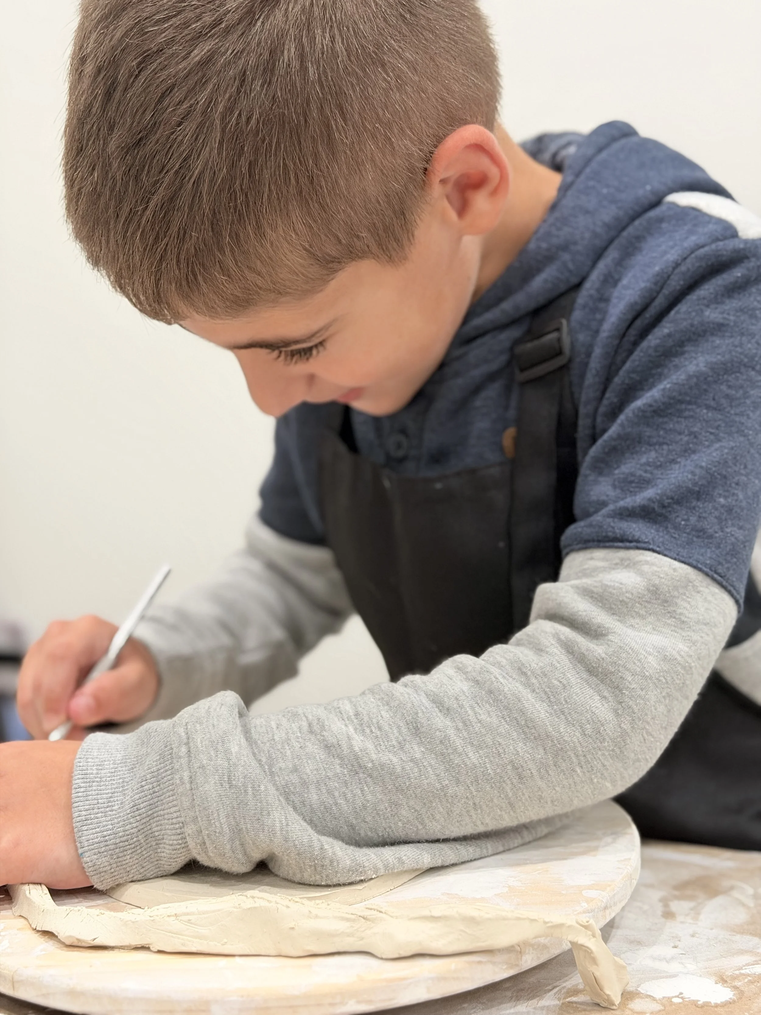 A young boy with short brown hair wearing a blue hoodie, working on a clay project with a roller on a wooden board.