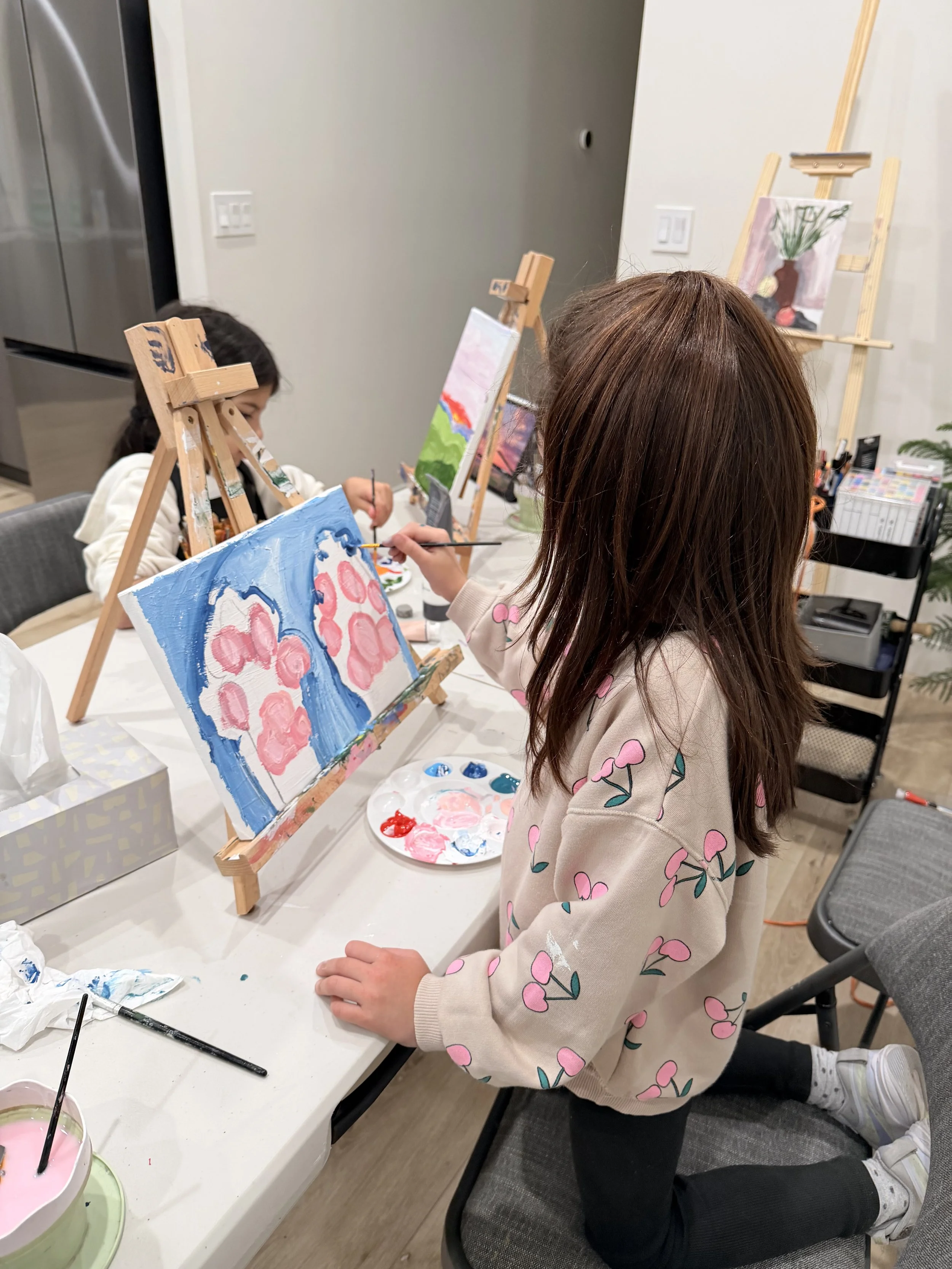A young girl with long brown hair, wearing a beige sweater with pink hearts and black pants, kneeling on a chair and painting on a canvas set on an easel at a table during an art activity in a room with paintings and art supplies.