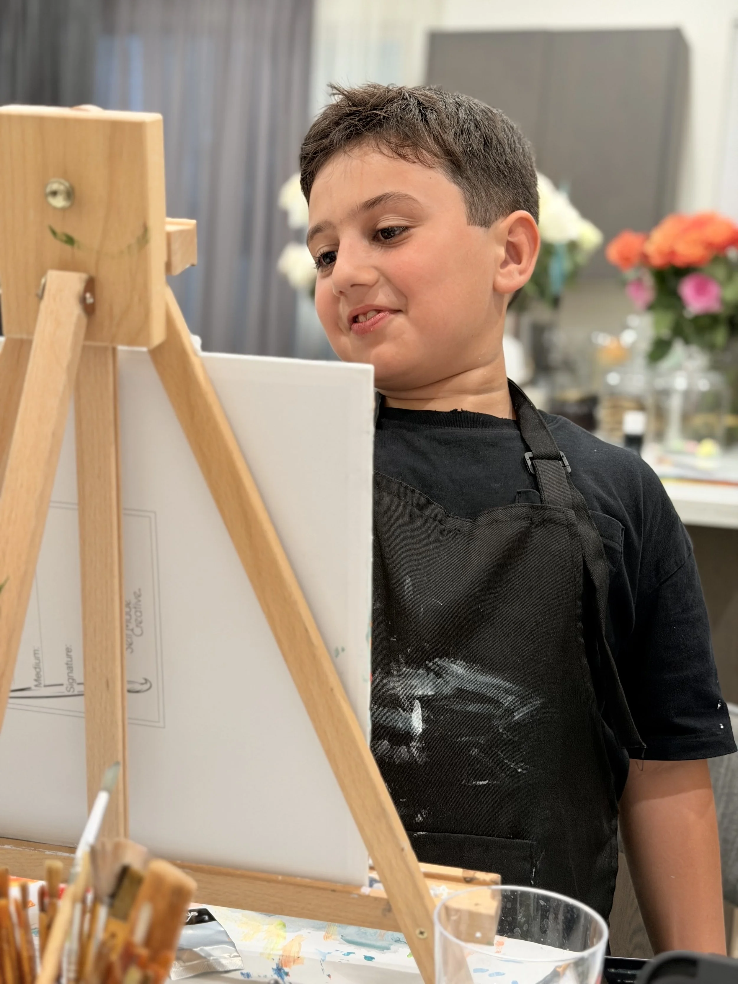 Young boy wearing a black apron painting on a canvas with an easel, with flowers in the background.