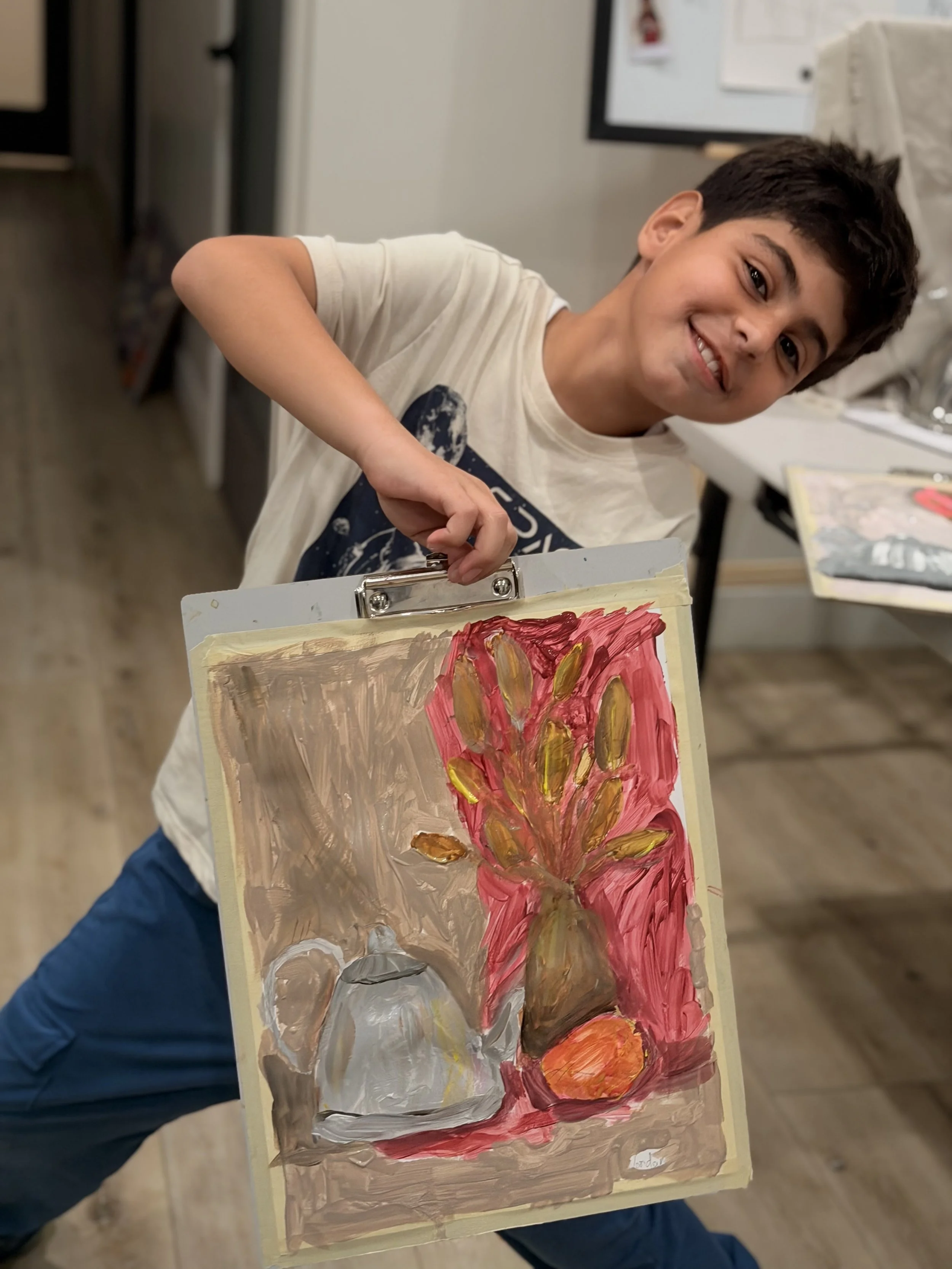Young boy smiling and holding a painting of a colorful flower in an art classroom.